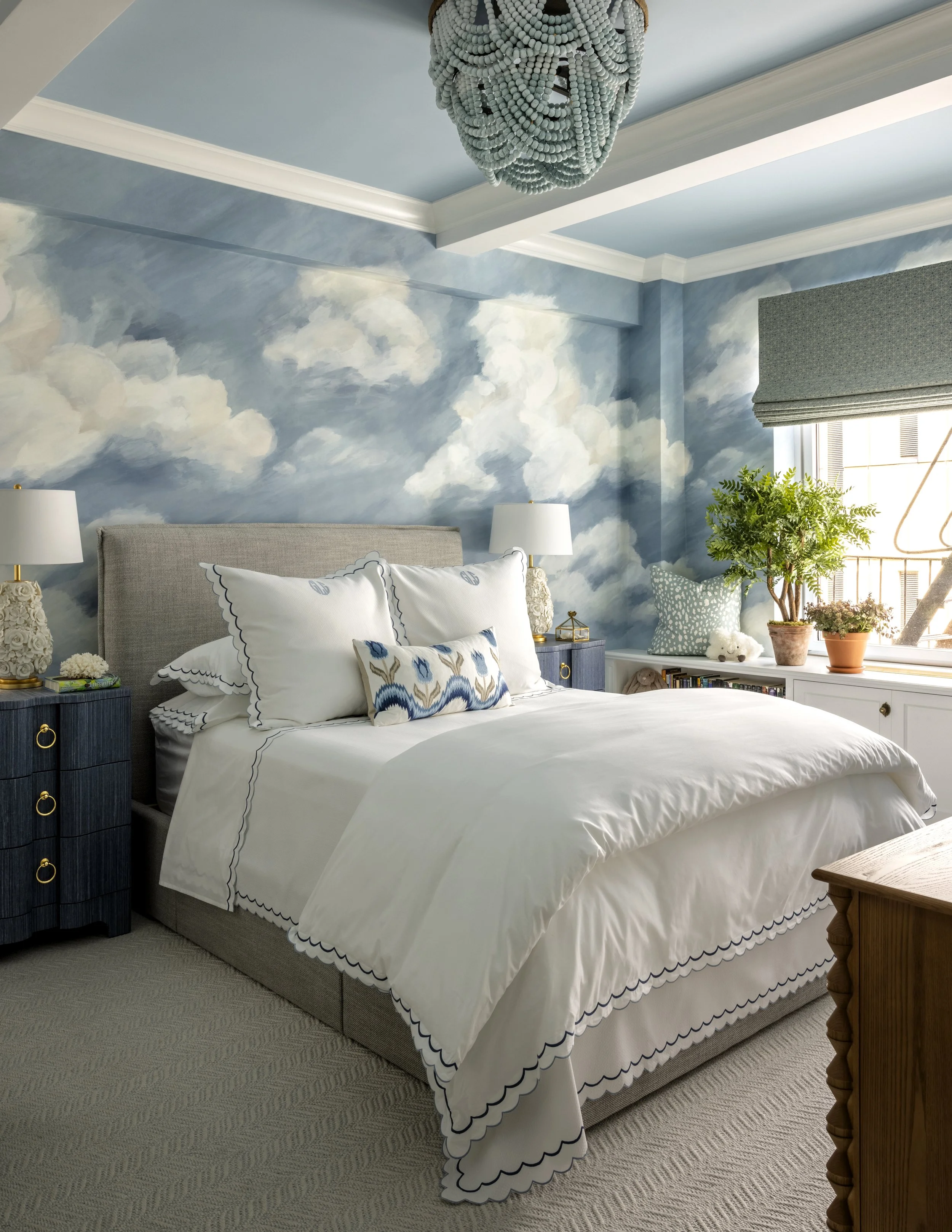 Bedroom with a sky-themed cloud mural on the wall, a bed with white and blue embroidered pillows, two bedside lamps, a window with a valance, and potted plants on a white windowsill.