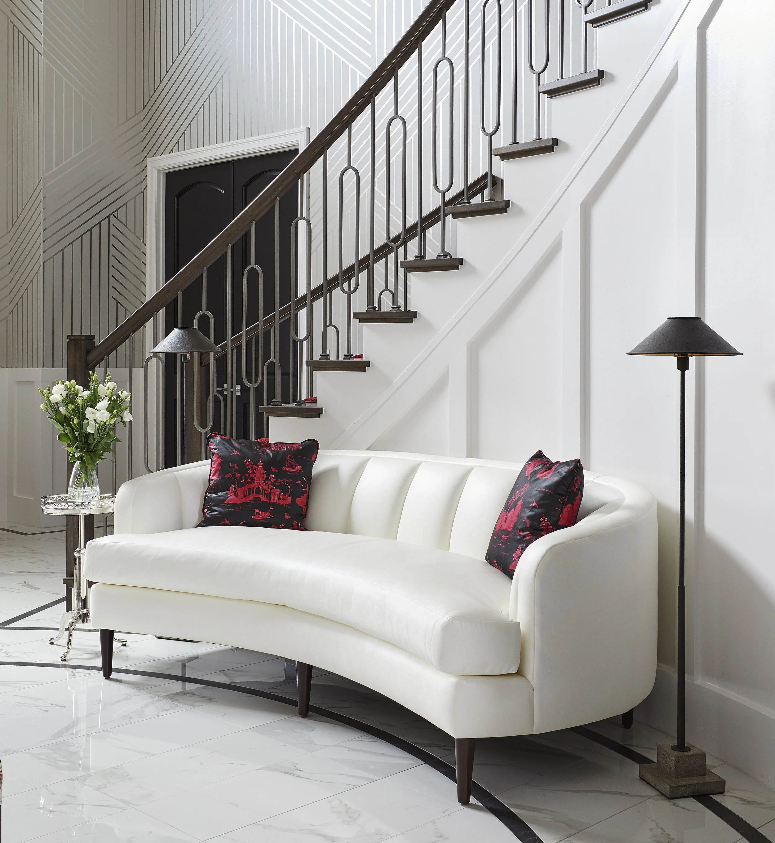 A modern living room with a white curved sofa, black and red decorative pillows, a glass side table with a bouquet of white flowers, a black floor lamp, and a staircase with black metal railing leading upstairs. The walls have a geometric patterned w