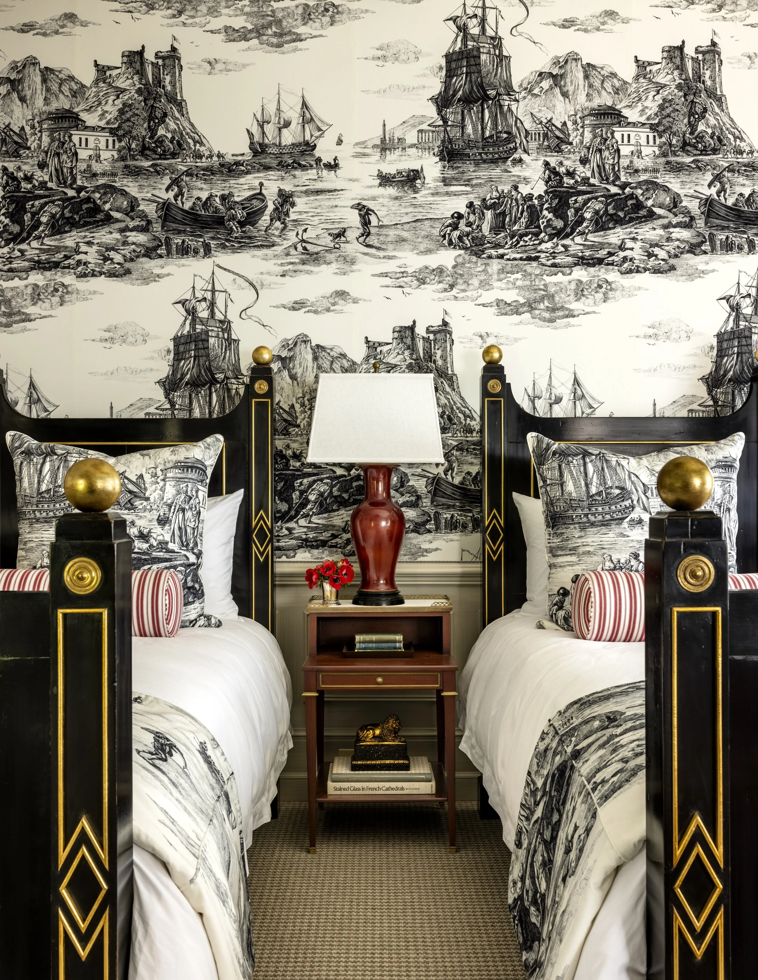 A bedroom with black and gold bed frames, white bedding, decorative pillows, and a detailed black-and-white mural of a historic harbor scene with ships, people, and castles.
