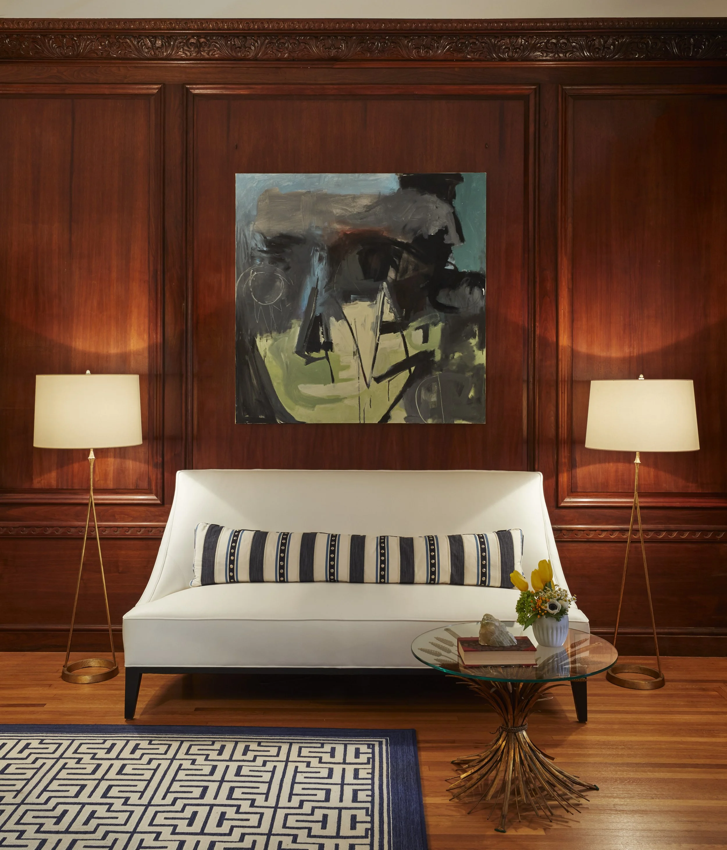 Living room with a white couch, black and white striped throw pillow, glass coffee table with a book, flower arrangement, and decorative rock, dark wood paneled walls with carved details, abstract painting, two tall floor lamps, and a patterned rug.