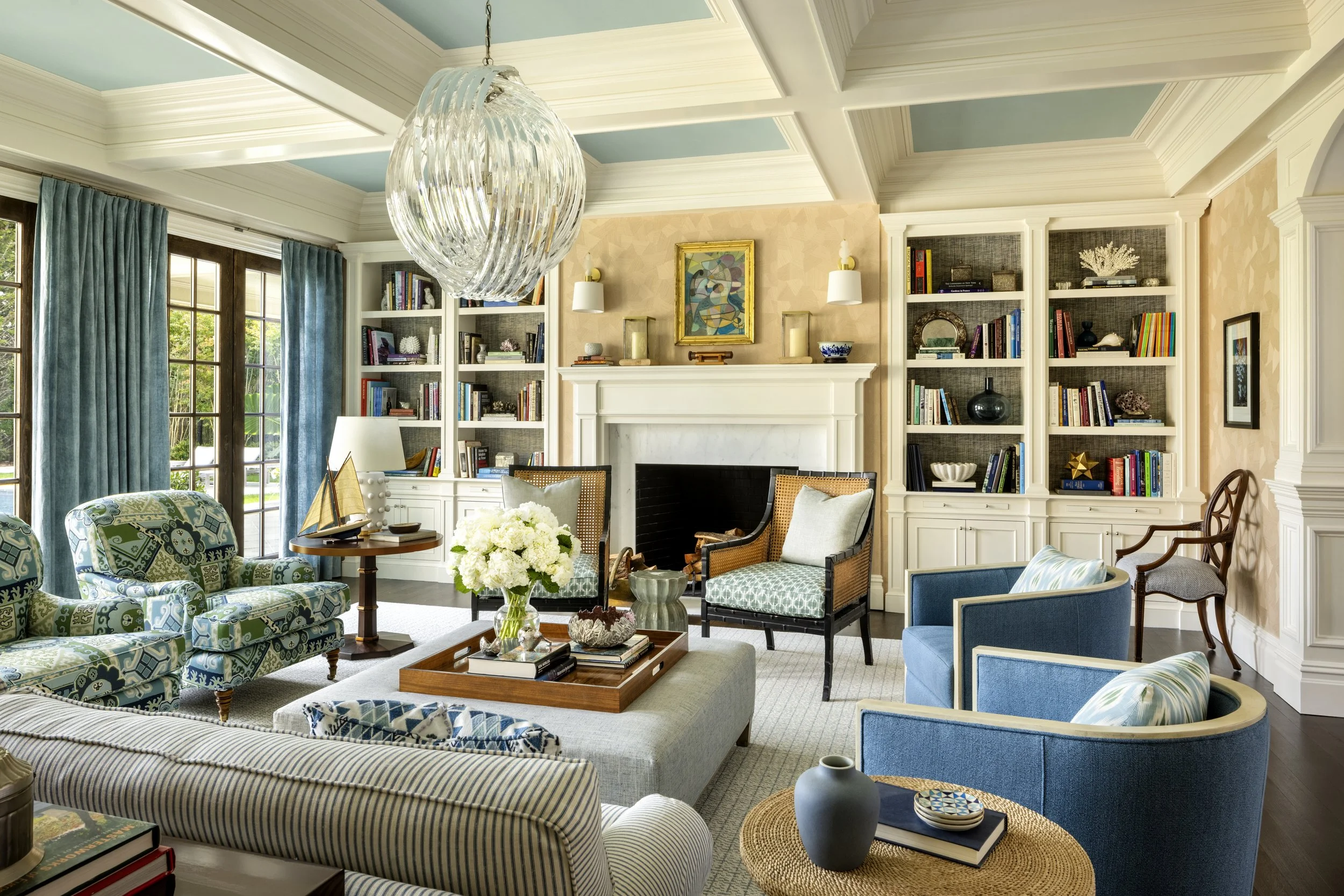 Living room with light blue ceiling, white molding, bookshelf, fireplace, and various armchairs. Large window with blue curtains, chandelier, and a table with a model sailboat and flowers.