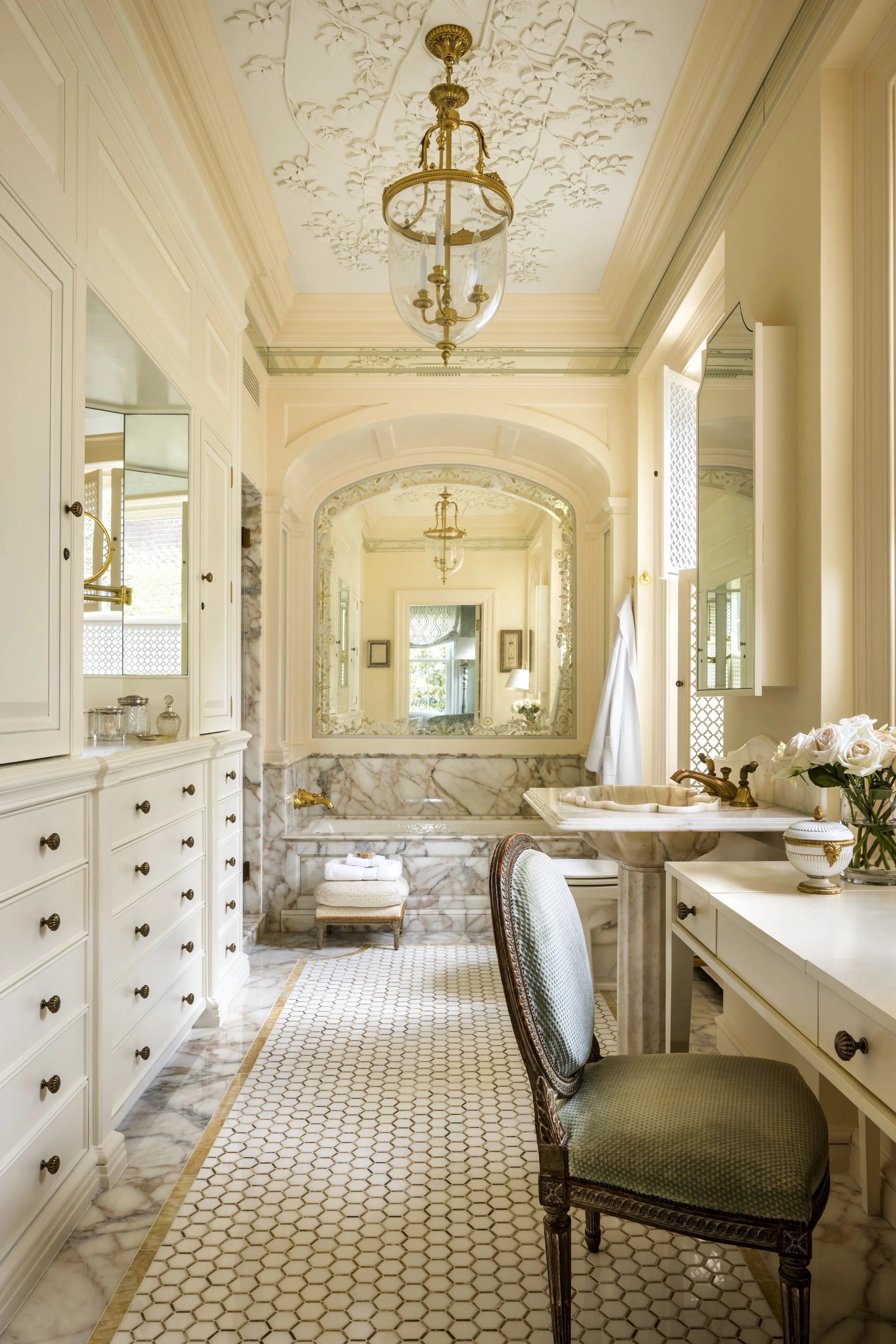 Elegant bathroom with cream and gold accents, featuring a marble bathtub, vintage chair, and large mirror, with intricate ceiling moldings and gold light fixtures.