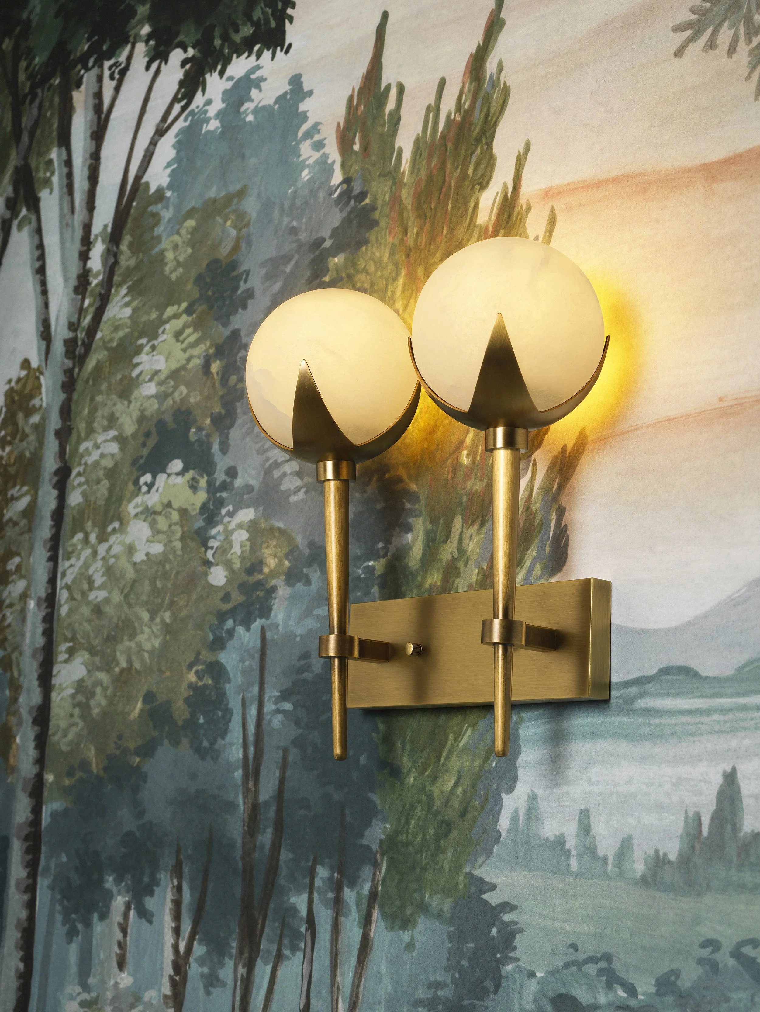 A vintage-style wall sconce with three spherical frosted glass lamps mounted on a wall with a landscape mural painting of trees and a cloudy sky.