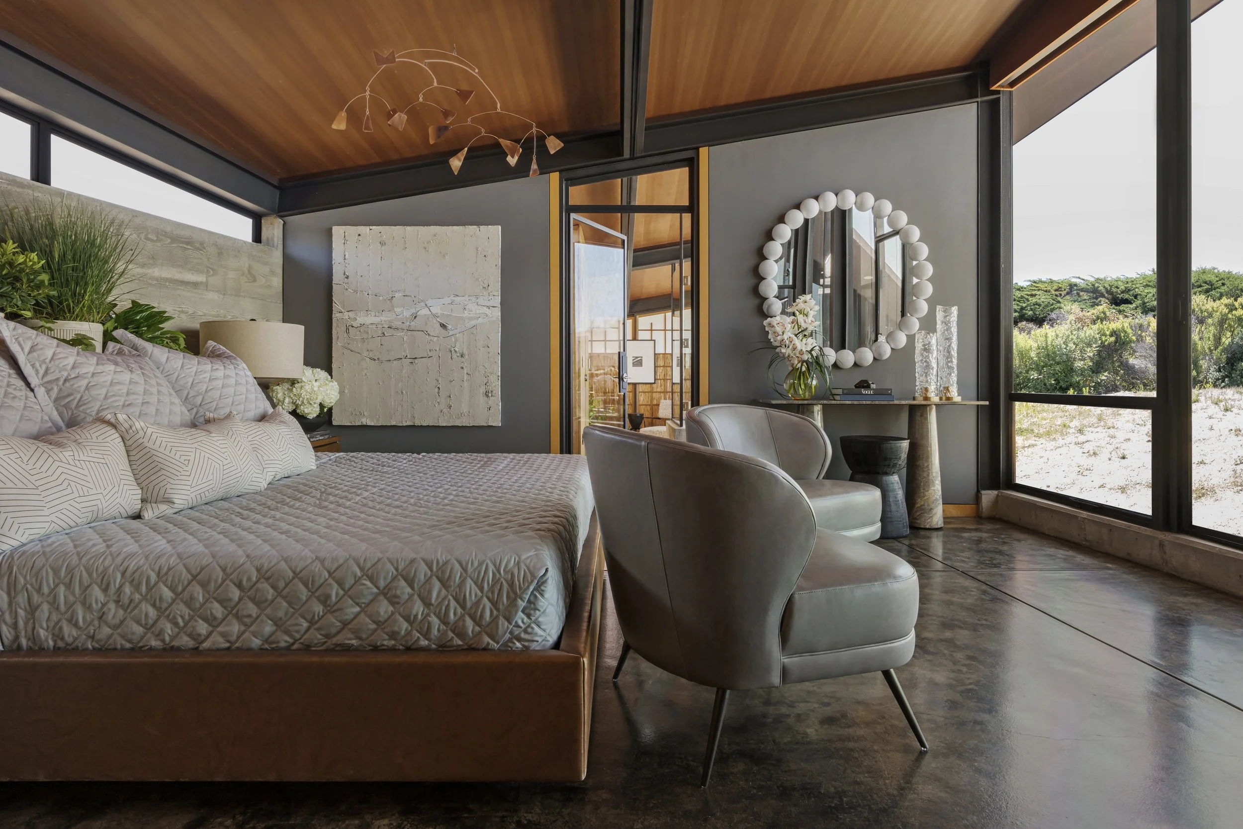 Modern bedroom with a large bed, a circular mirror with white lights around it, stylish chairs, decorative flowers, large windows with a view of greenery, and a wood ceiling.