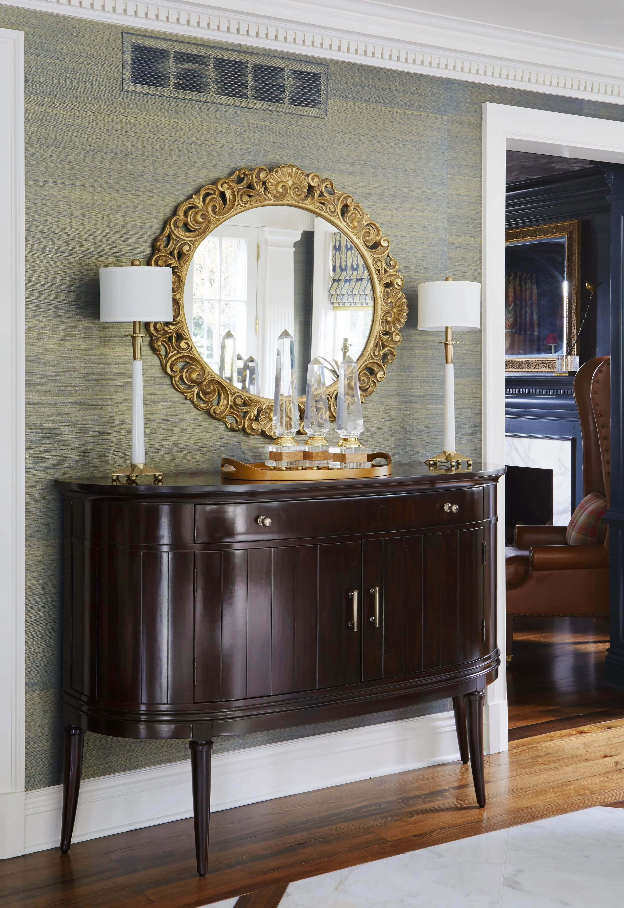 A dark wood sideboard with a curved front, standing against a textured green wall. On top of the sideboard, there are three tall, clear glass objects on a wooden tray. Flanking the sideboard, there are two white lamps with gold bases. A gold-framed d