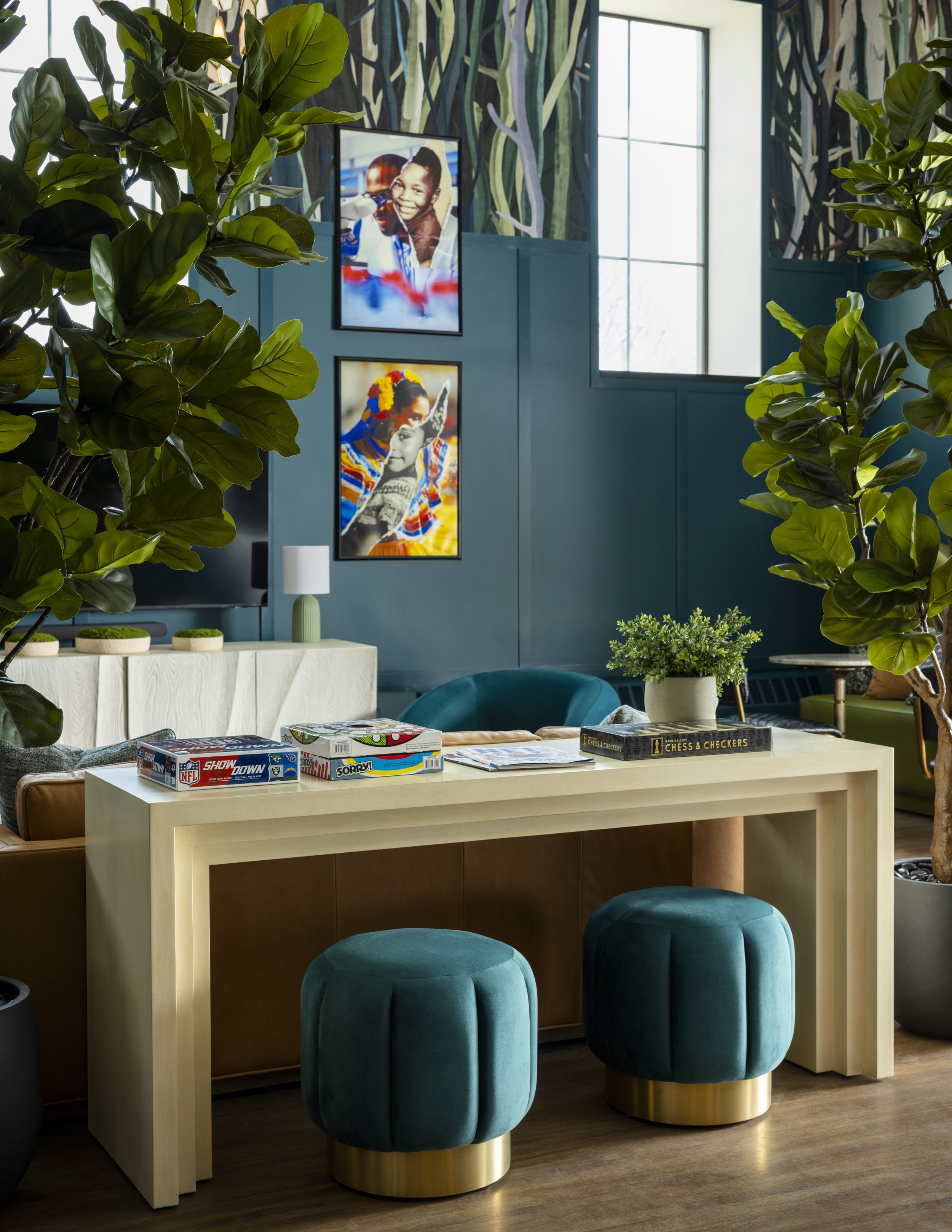 A modern living room with a beige table, teal velvet ottomans with gold bases, large green indoor plants, a white sideboard, colorful framed photographs of children and cultural scenes, a wall mural depicting trees, and a window letting in natural li