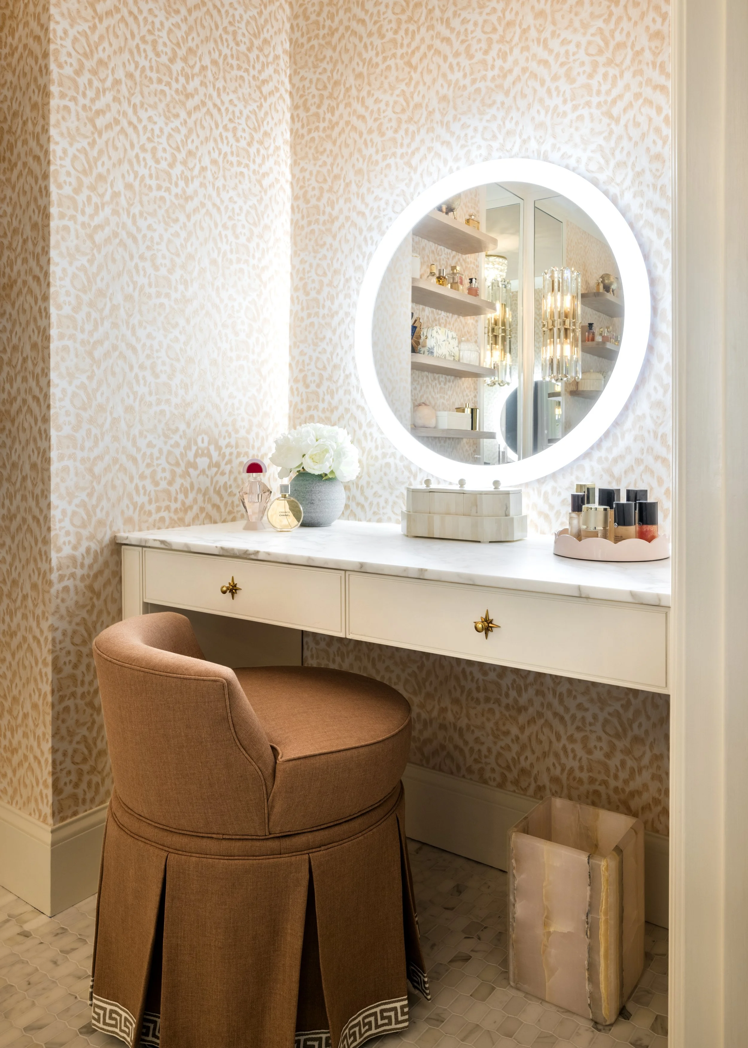A vanity area with a round illuminated mirror, pink animal print wallpaper, brown upholstered chair, marble top dresser with decorative items, flowers, perfume, and makeup. Shelves with bottles and decorative objects are visible in the mirror's refle