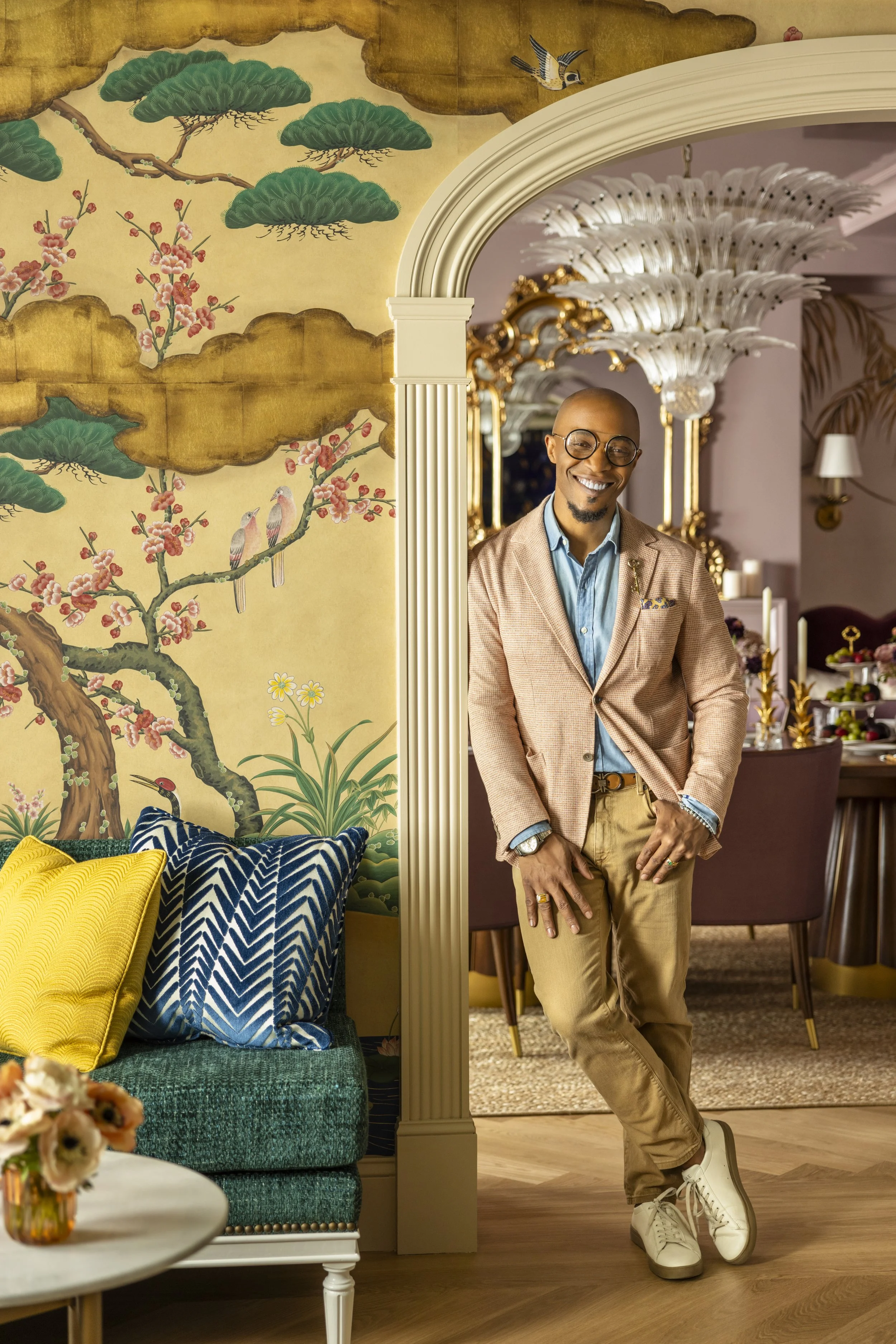 Corey Damen Jenkins wearing glasses, a beige blazer, and khaki pants, standing in a stylish, vintage-inspired room with ornate lighting fixtures, a mural with Asian-inspired art, and a cozy sitting area with colorful pillows.