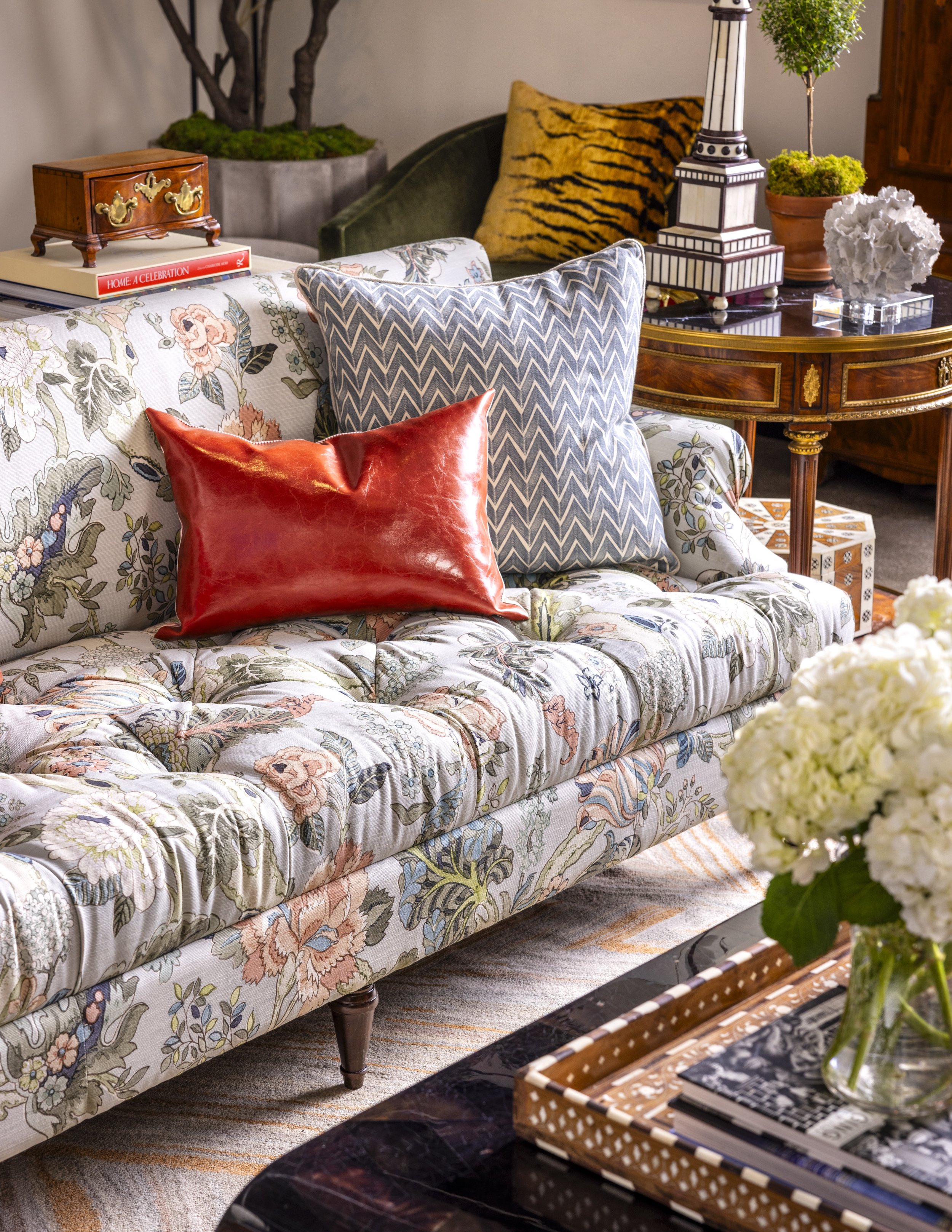 Living room seating area with a floral upholstered sofa, decorative pillows, a wooden side table with books and decorative items, and a round wooden table with a lamp, plants, and decor. There is a white flower arrangement in a glass vase in the fore