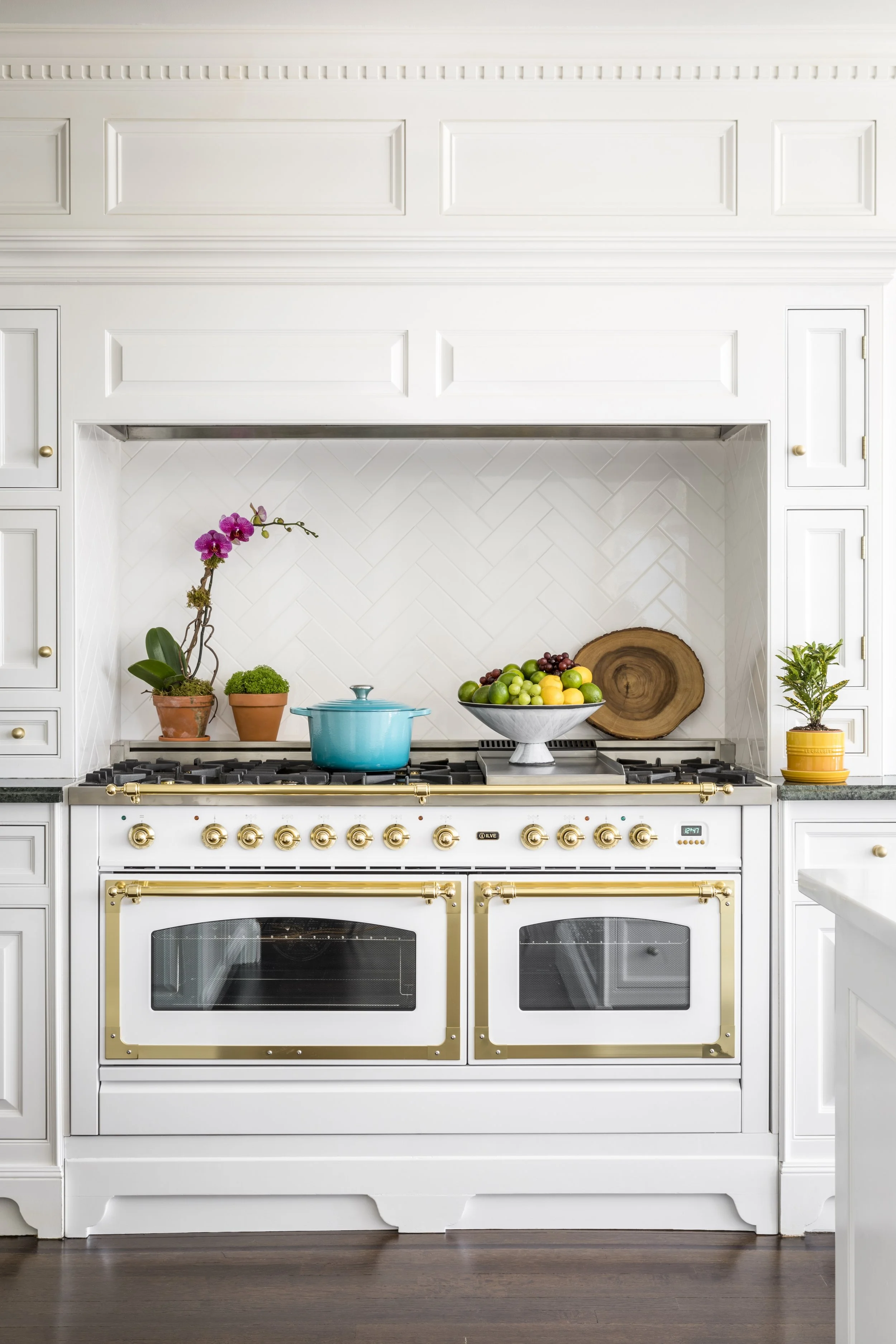 White kitchen stove with gold accents, plants, a bowl of fruits, and a teal pot on top.