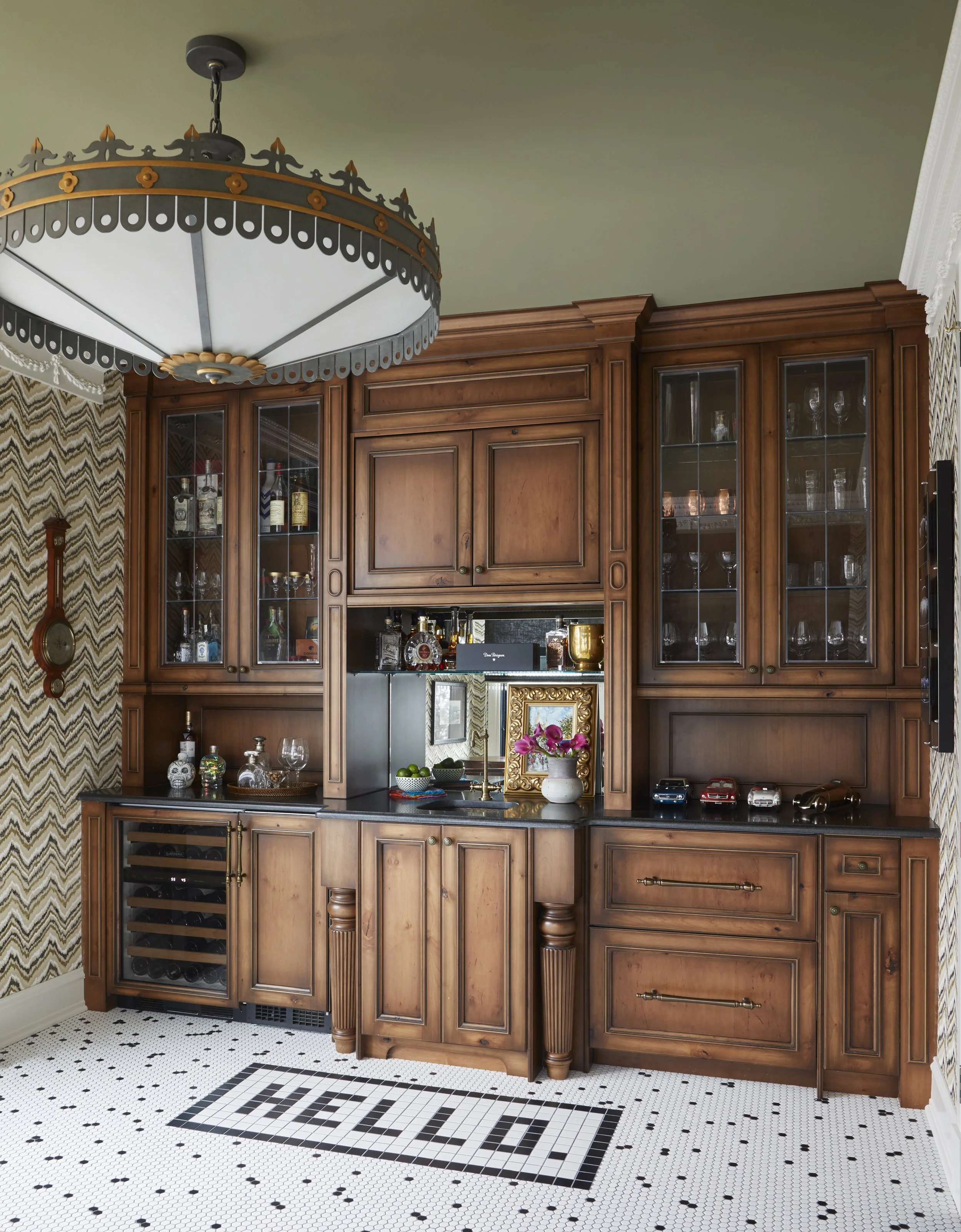 Traditional wooden bar cabinet with glass doors filled with liquor bottles and glassware, with a black countertop, set against vintage wallpaper with a champagne gold and cream chevron pattern, and decorated with pink flowers and small toy cars on th