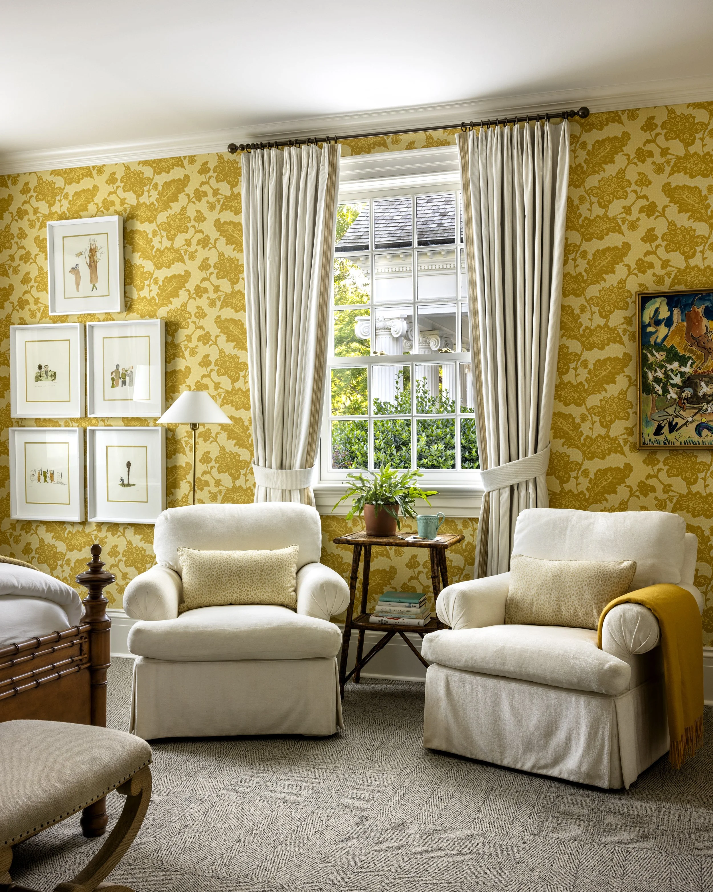 A cozy living room with cream-colored armchairs, a wooden side table with a potted plant, and a large window with cream curtains, yellow floral wallpaper, and framed artwork on the wall.