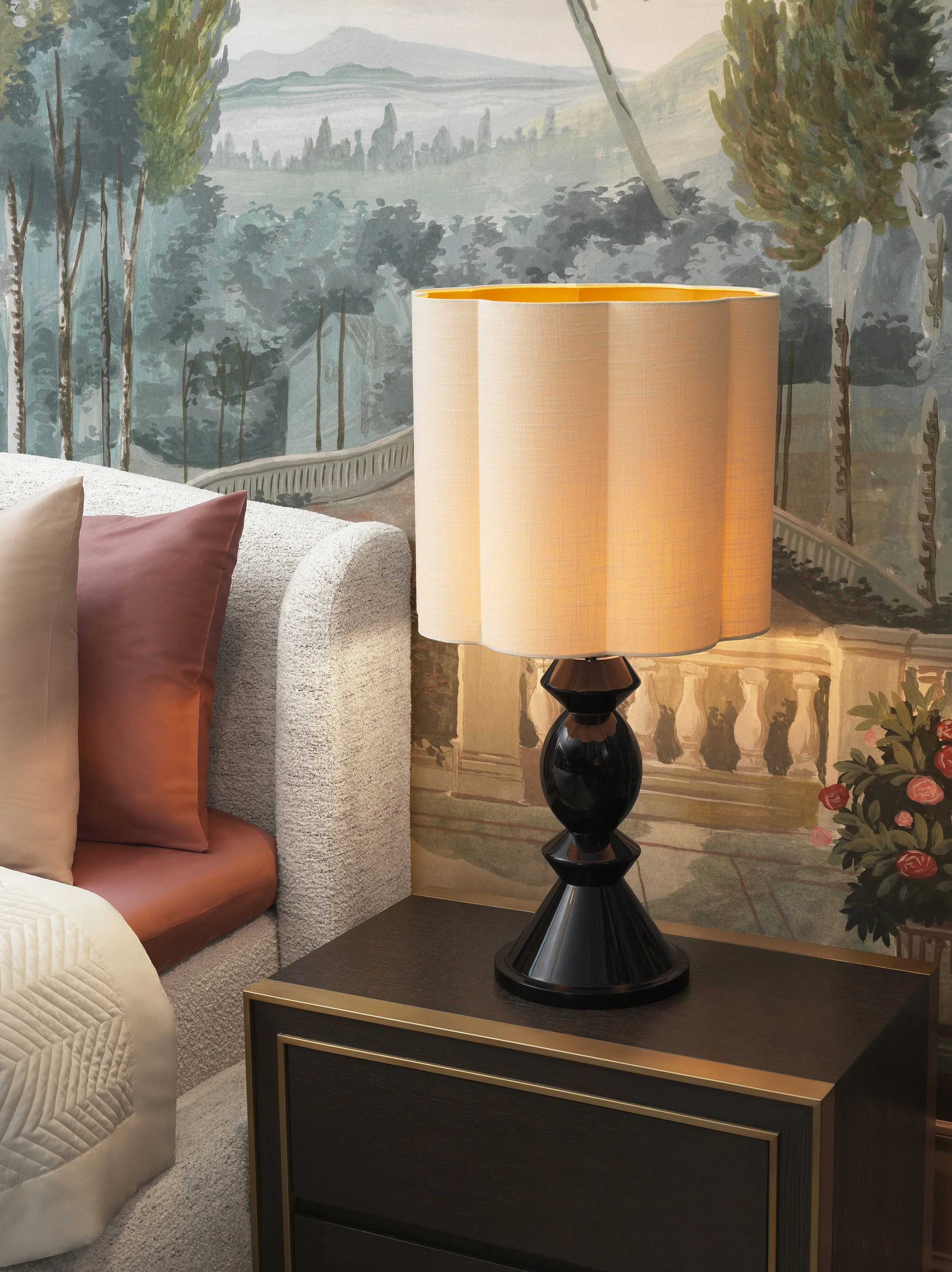 A room with a bed and a wall mural featuring a scenic landscape of trees and mountains. A black and gold side table holds a large beige table lamp with a black base. Part of a beige couch with a pink pillow is visible on the left.