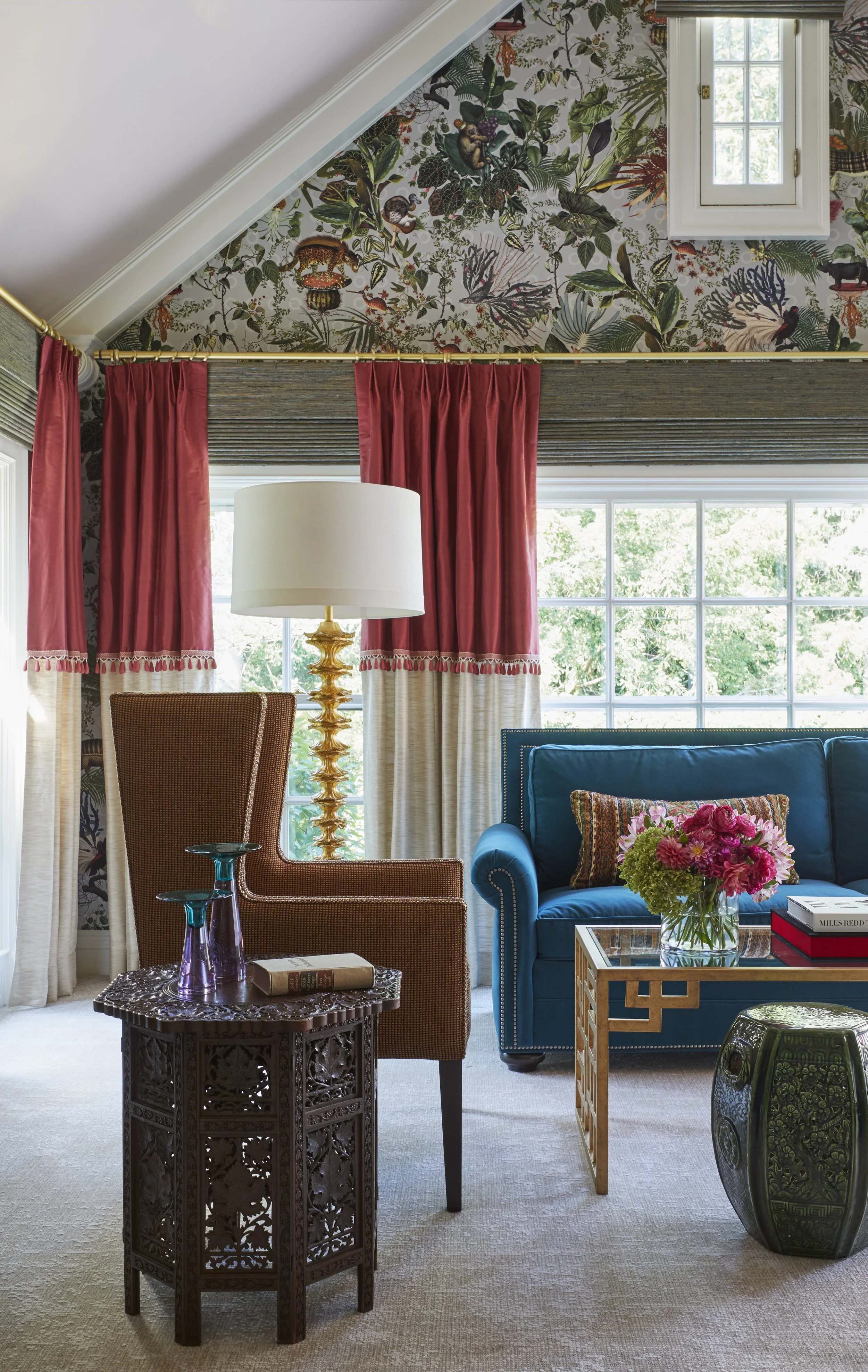 A cozy living room with a large window showing greenery outside. There are colorful velvet curtains, a blue sofa with a floral pillow, a rattan armchair, a glass-top coffee table with books and a flower arrangement, a carved wooden side table with gl