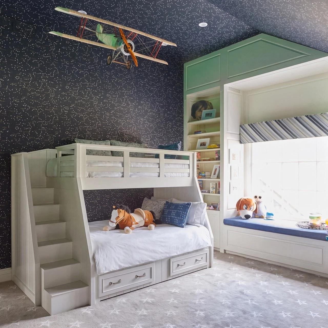 Children's bedroom with starry night sky wallpaper, a white bunk bed with a staircase, plush toys including a horse and a dog on the bed, built-in bookshelves filled with books, a window seat with stuffed animals, and a ceiling-mounted vintage airpla