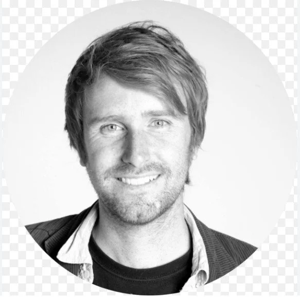 Black and white portrait of a smiling man with medium-length hair, wearing a blazer over a collared shirt
