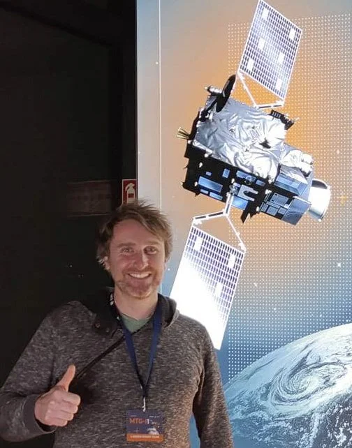 A smiling man with a thumbs-up gesture standing in front of an illustration of a satellite orbiting Earth at an exhibition or science center.