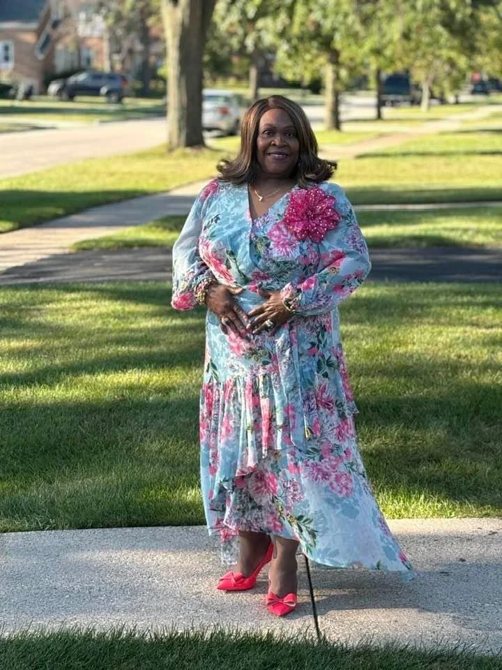 A woman standing on a sidewalk in a park, wearing a floral dress with pink accents, pink heels, and smiling at the camera.