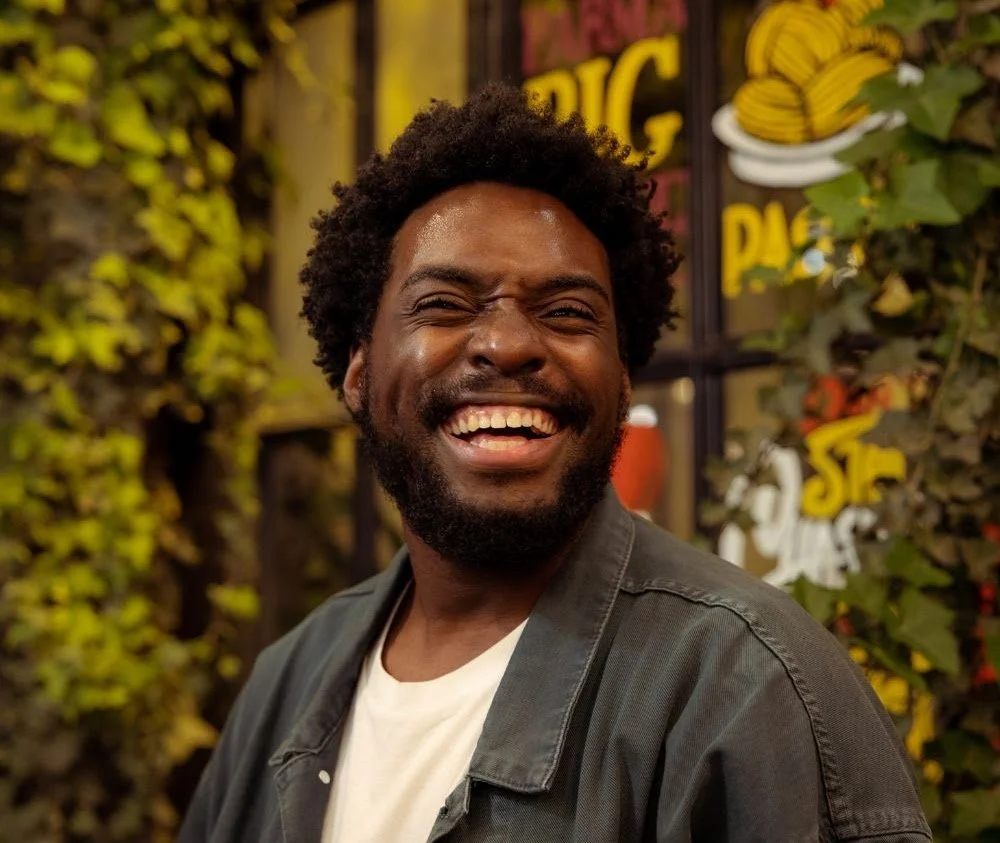 A man with a curly afro and beard, smiling and laughing, standing in front of a decorated background with illustrations and text.
