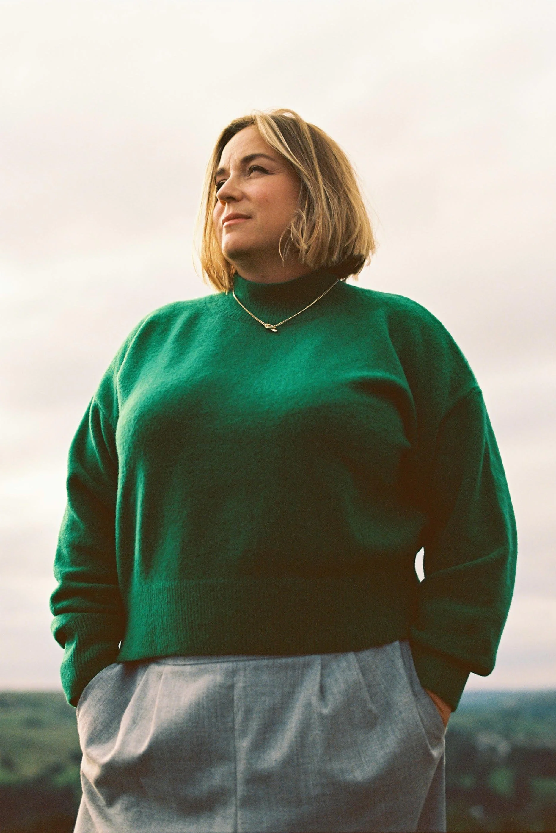 Penny Lee, niche brand strategist and founder of The Brand Directoress, standing outdoors in a green turtleneck jumper and grey trousers, hands in pockets, looking upward with quiet confidence against a soft overcast sky.