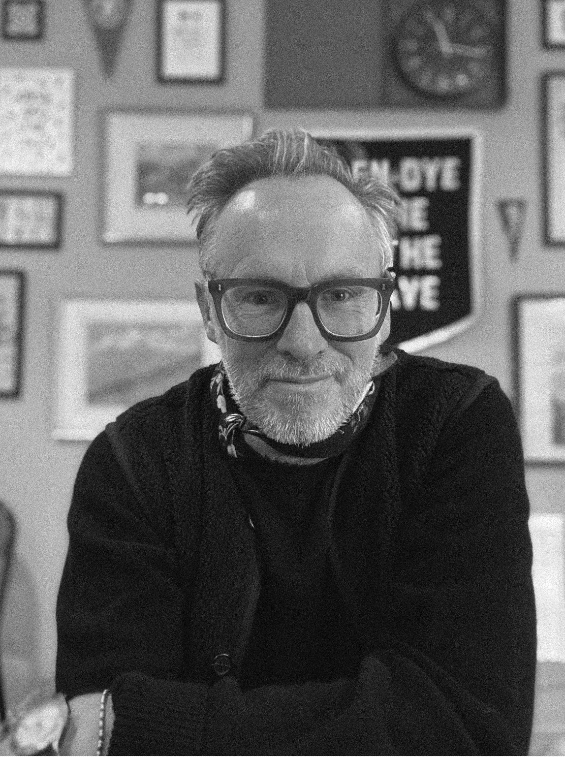 A man with glasses and a beard smiling in a room with a gallery wall of framed pictures and posters behind him.