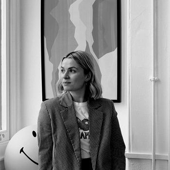 A woman with shoulder-length hair wearing a blazer stands next to a large smiling balloon in front of an abstract painting in a room.