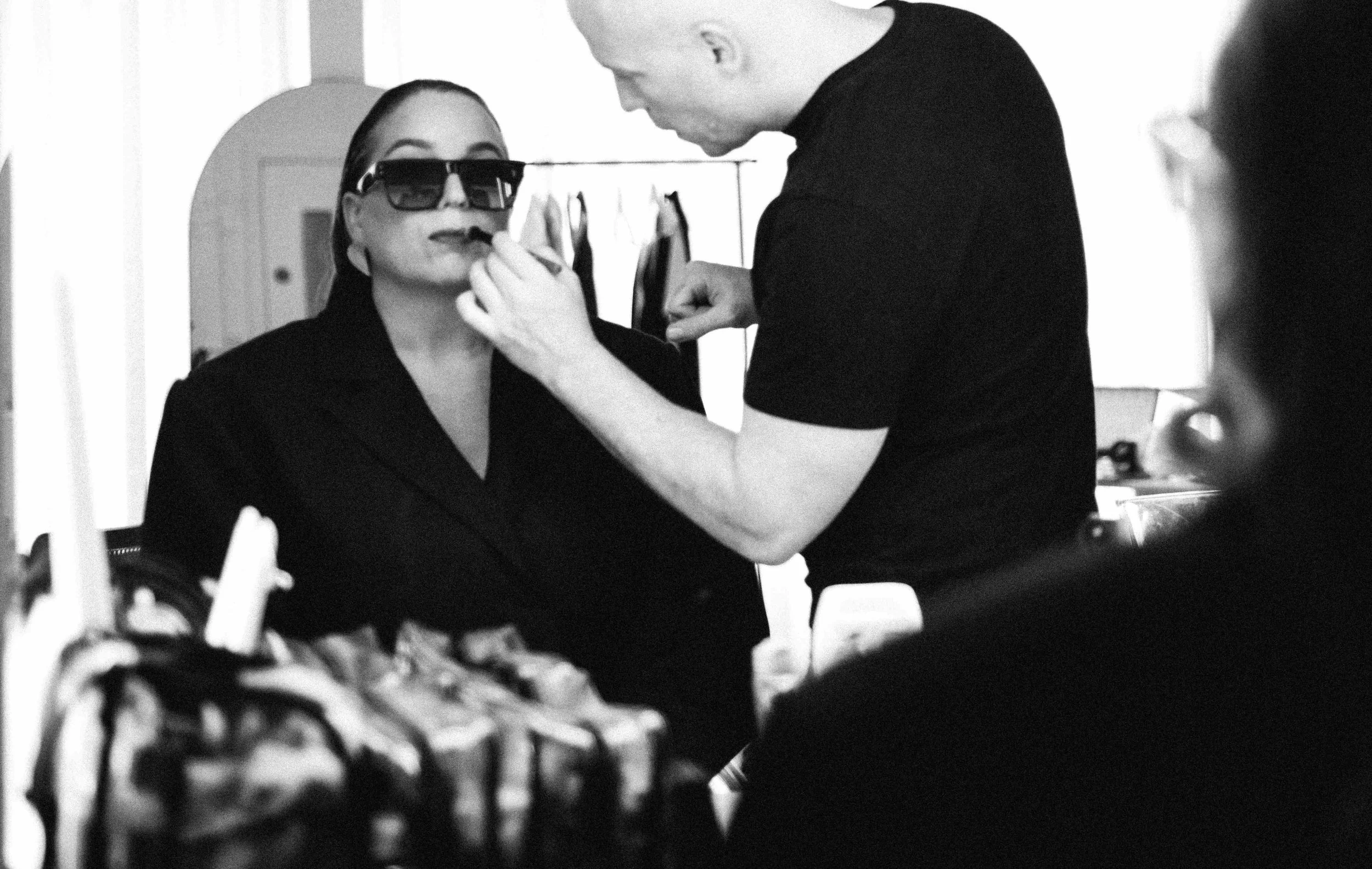 Penny Lee wearing sunglasses and a black suit has makeup applied by a man, who is standing close to her with a focused expression. The scene is in a makeup or dressing room.
