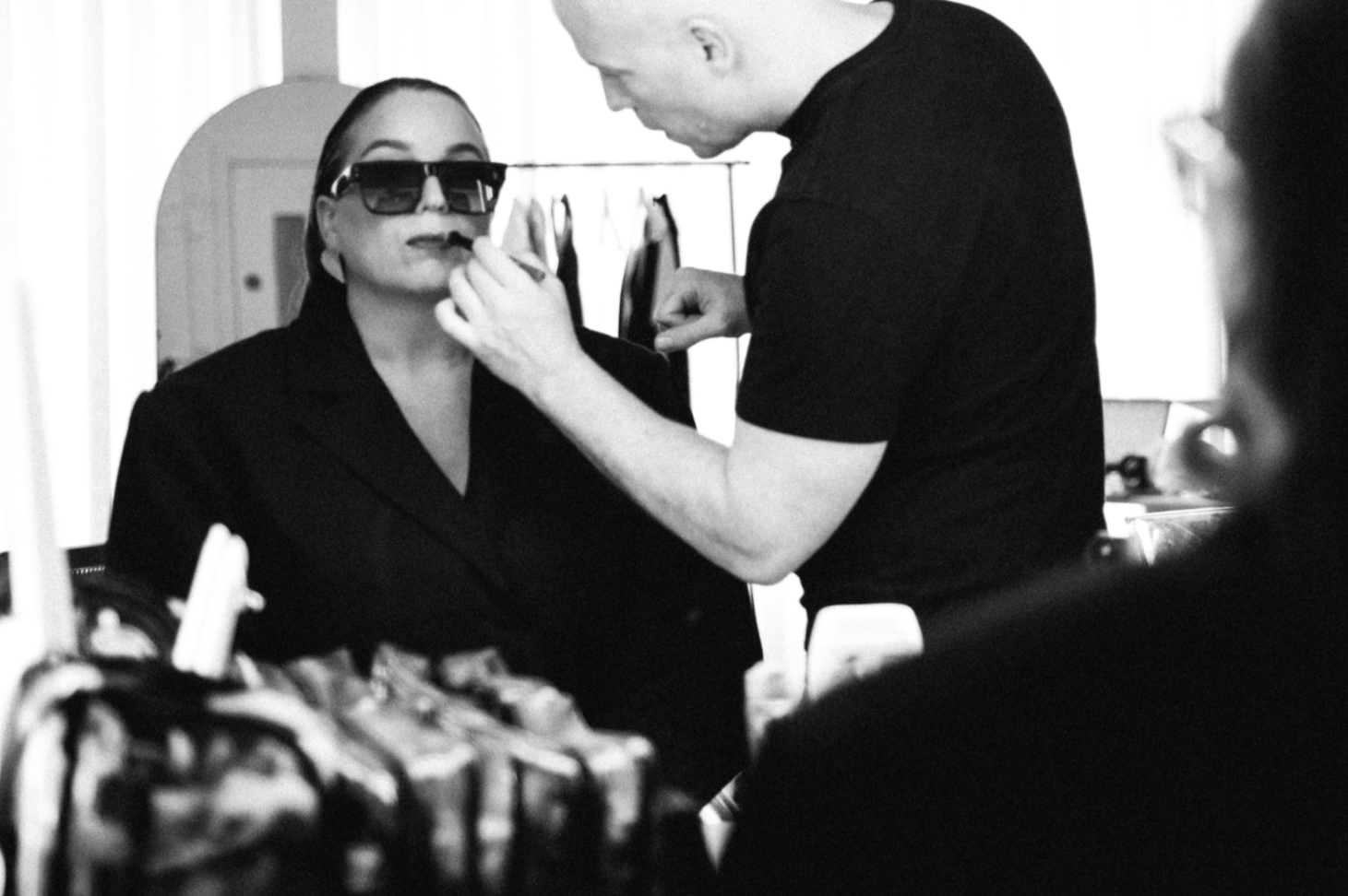 A fashion stylist applies makeup or face paint to a woman wearing large sunglasses, as she prepares for this photoshoot.