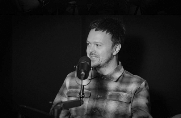A man with dark hair and a beard, wearing a plaid shirt, speaking into a microphone in a recording studio.