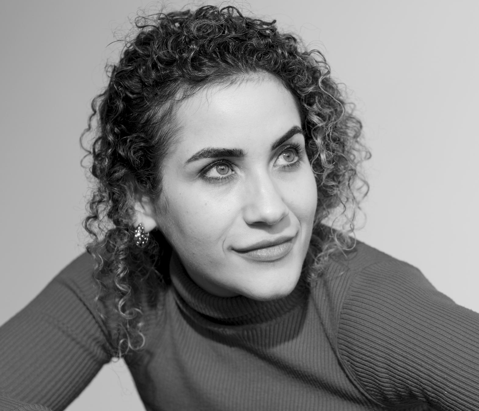 A black and white portrait of a woman with curly hair, wearing earrings and a ribbed, long-sleeve top, looking off to her right with a thoughtful expression.