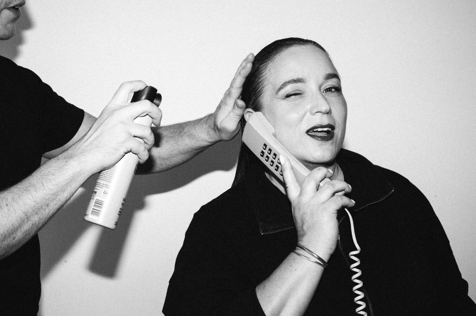 A woman with dark hair and dark lipstick is talking on a corded landline telephone while a person sprays hairspray on her hair. The woman is winking and smiling, and the scene is in black and white.