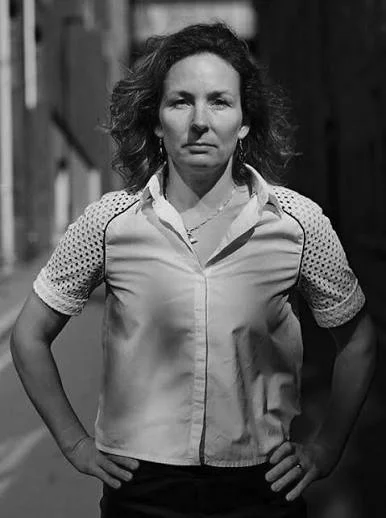 A woman with curly hair standing with her hands on her hips, wearing a short-sleeved shirt, outdoors with a wooden structure in the background.