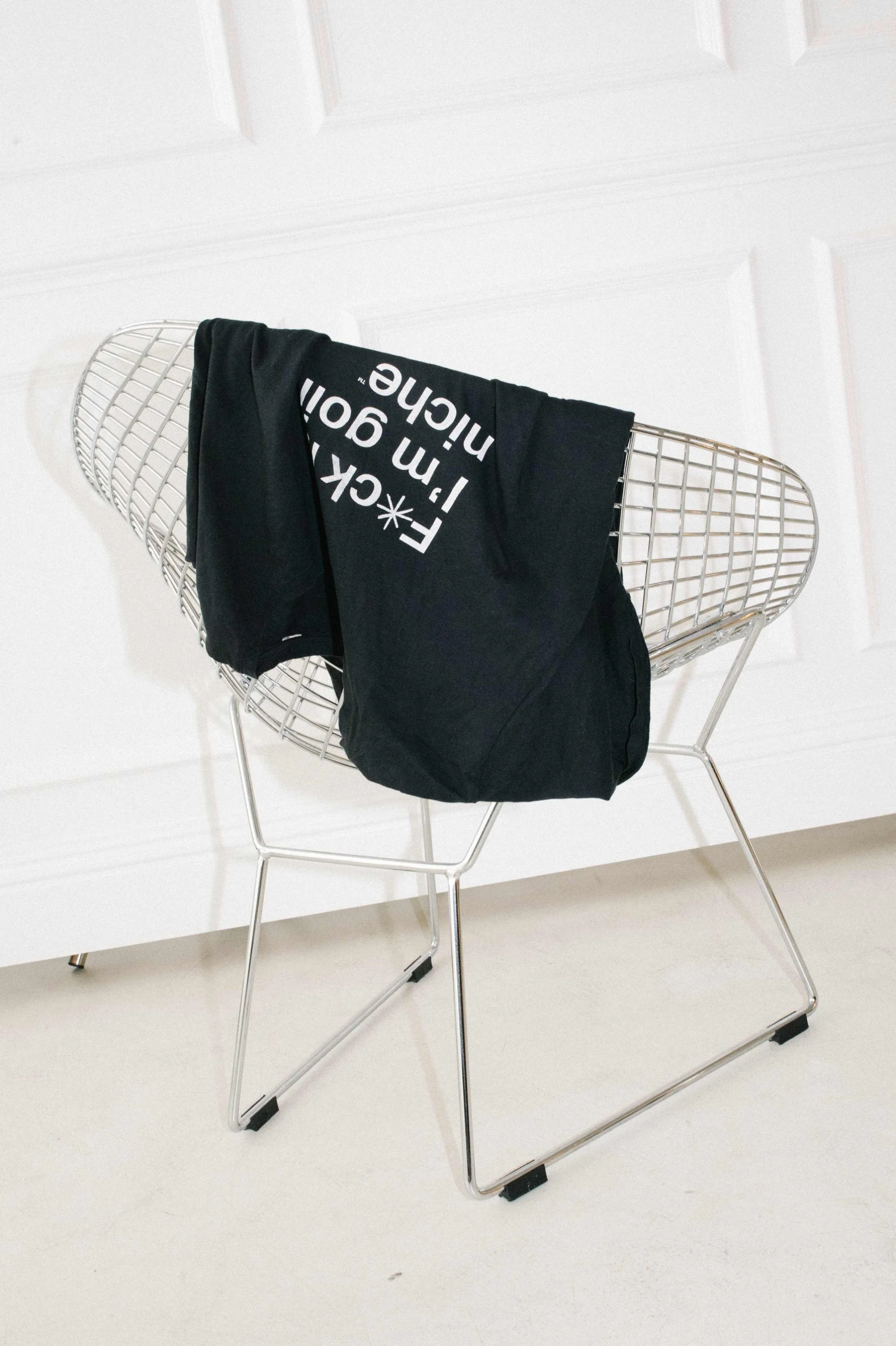 A wire chair with a black t-shirt draped over it with white text reading F*ck it I'm going niche, against a plain white wall.