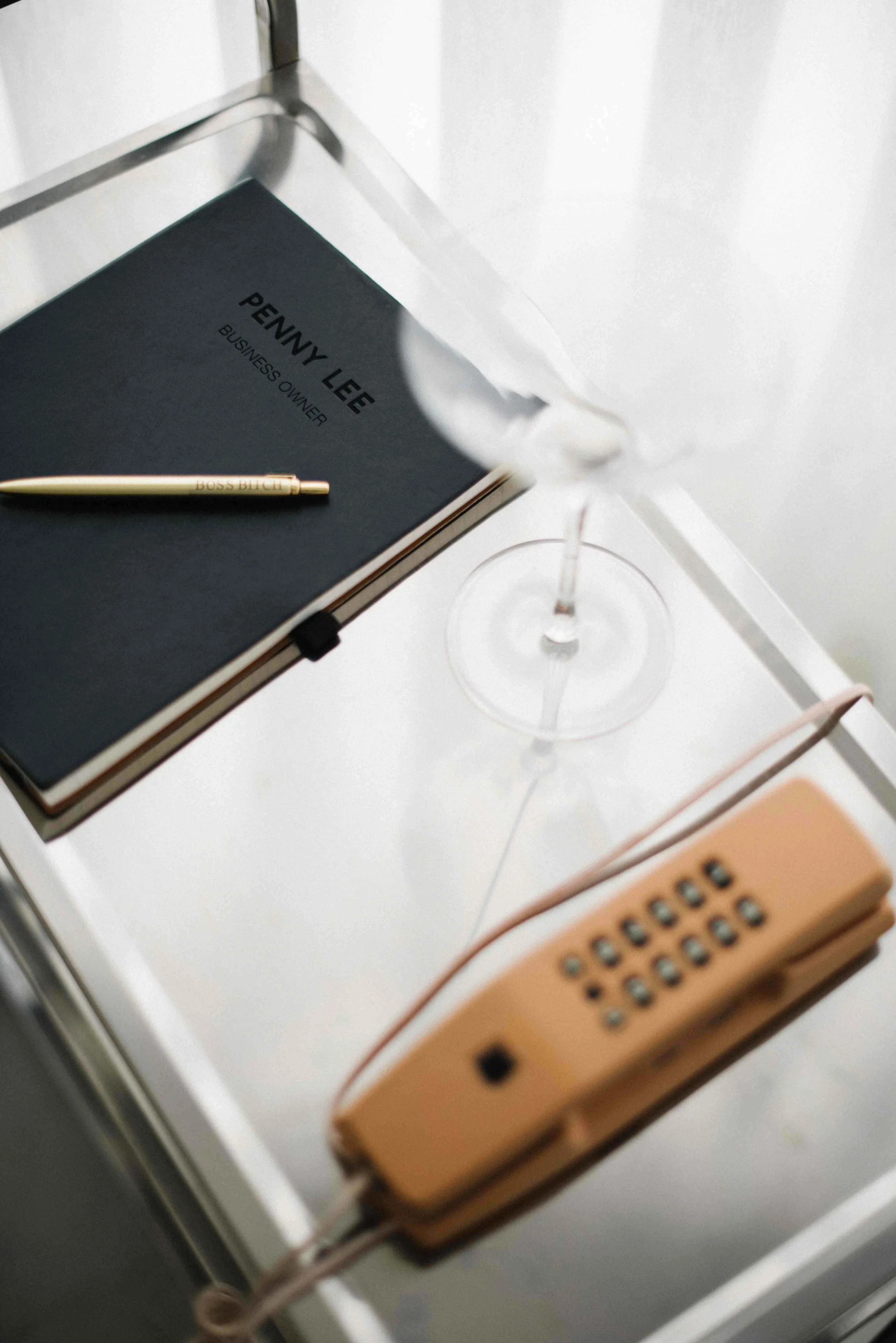 A gold-colored pen with the words 'BOSS BITCH' engraved on it, resting on a black notebook labelled 'PENNY LEE BUSINESS OWNER', placed on a white surface next to a cocktail glass and a retro peach telephone.