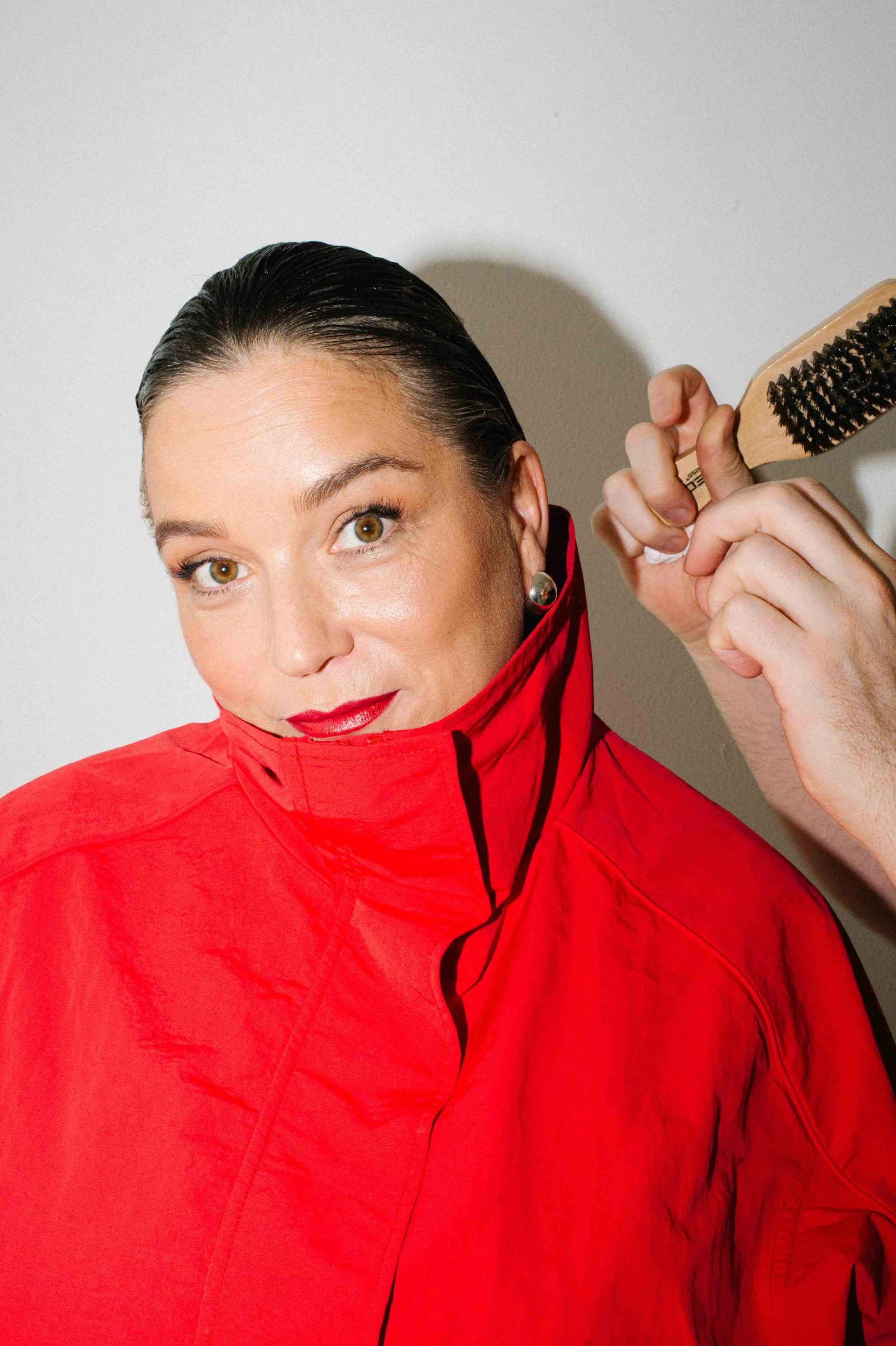 Penny Lee with slicked-back dark hair and light skin, wearing a red jacket, stylist hand holding a hairbrush near her head, standing against a plain white background.