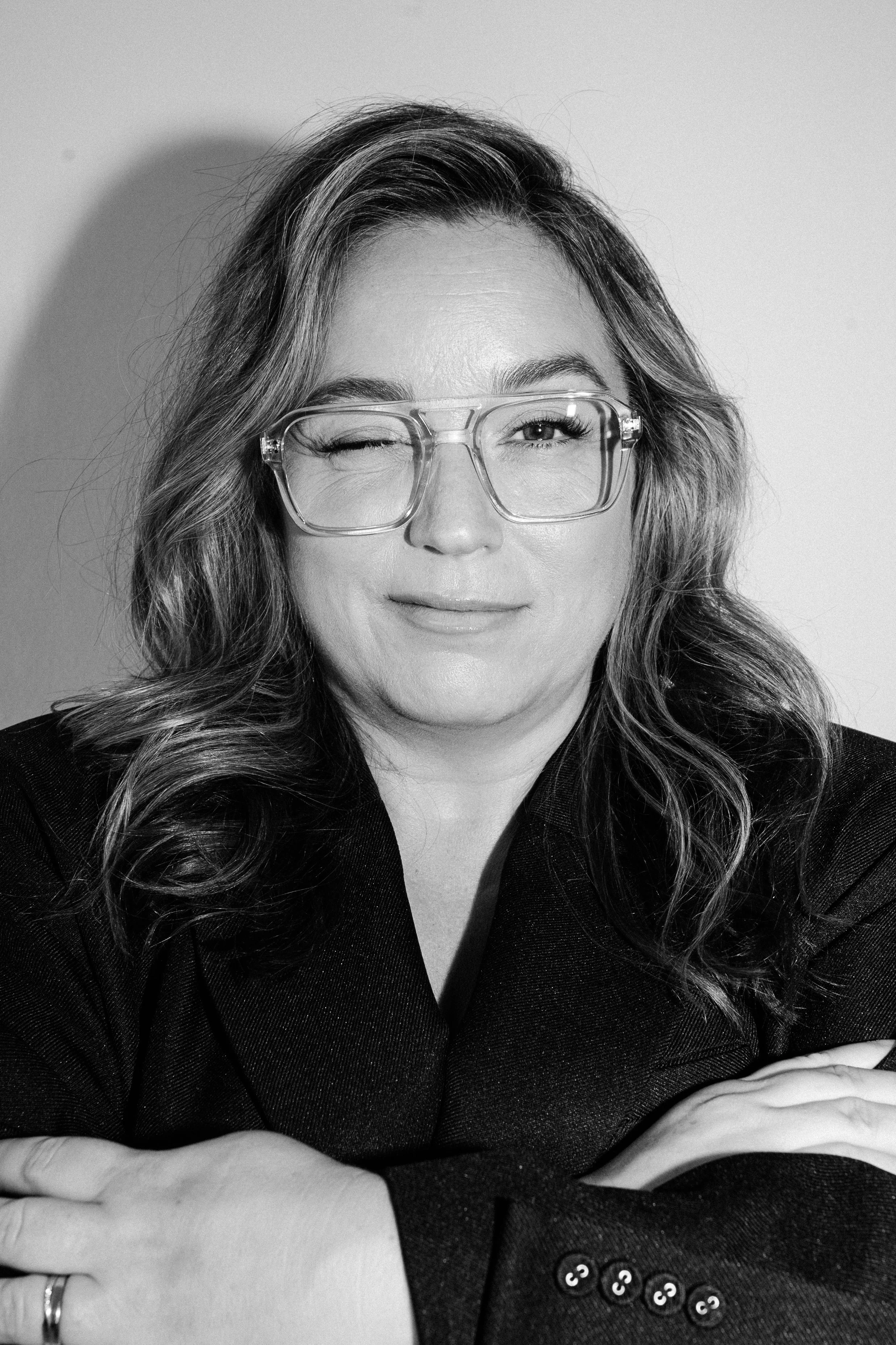 A woman with wavy hair, wearing glasses, winking and smiling. She is dressed in a dark blazer with buttons and has her arms crossed in front of her in bold stance.