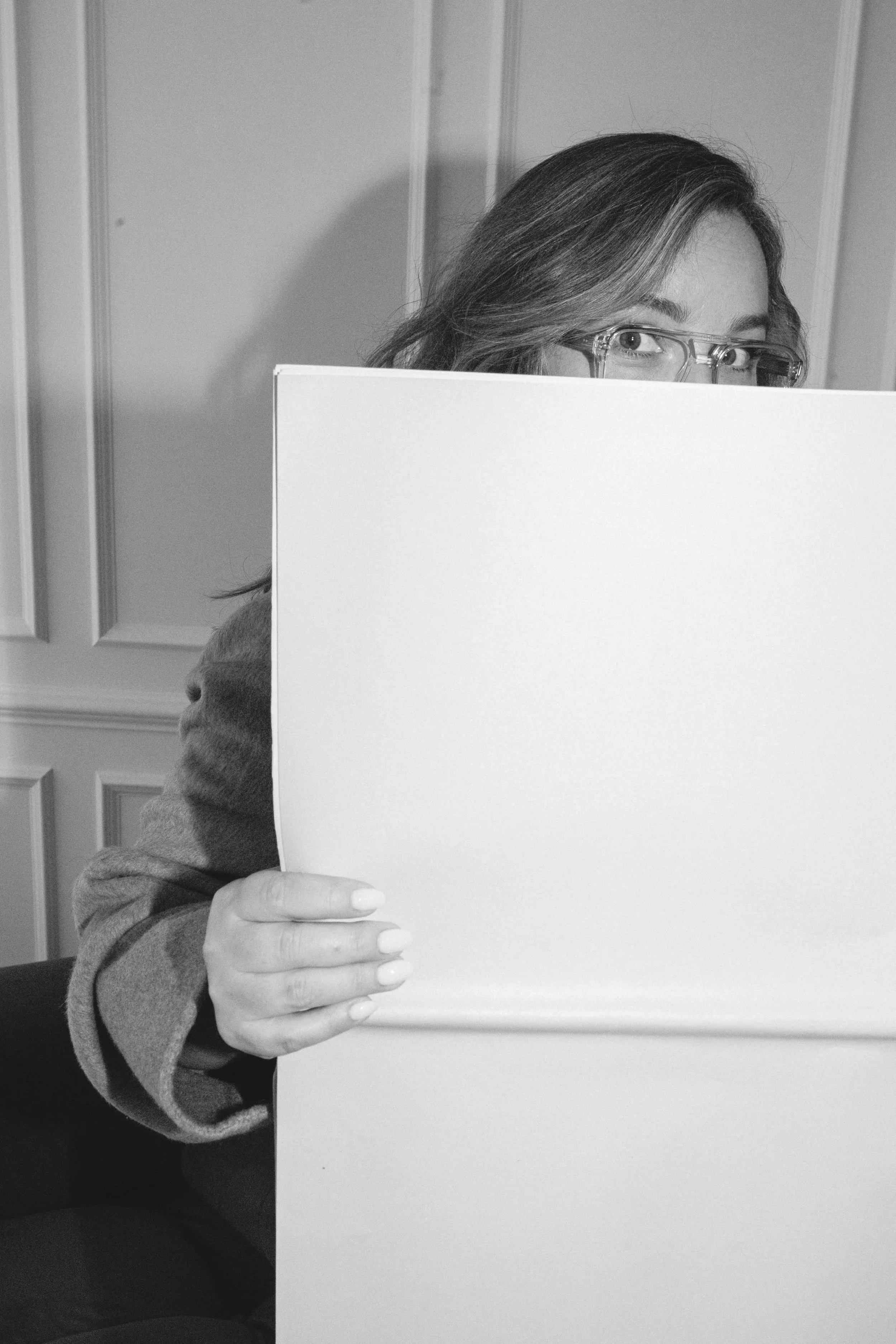 Black and white photo of Penny Lee with glasses holding a blank large white poster board in front of her face, covering her body from the shoulders down, with wooden panel walls in the background.