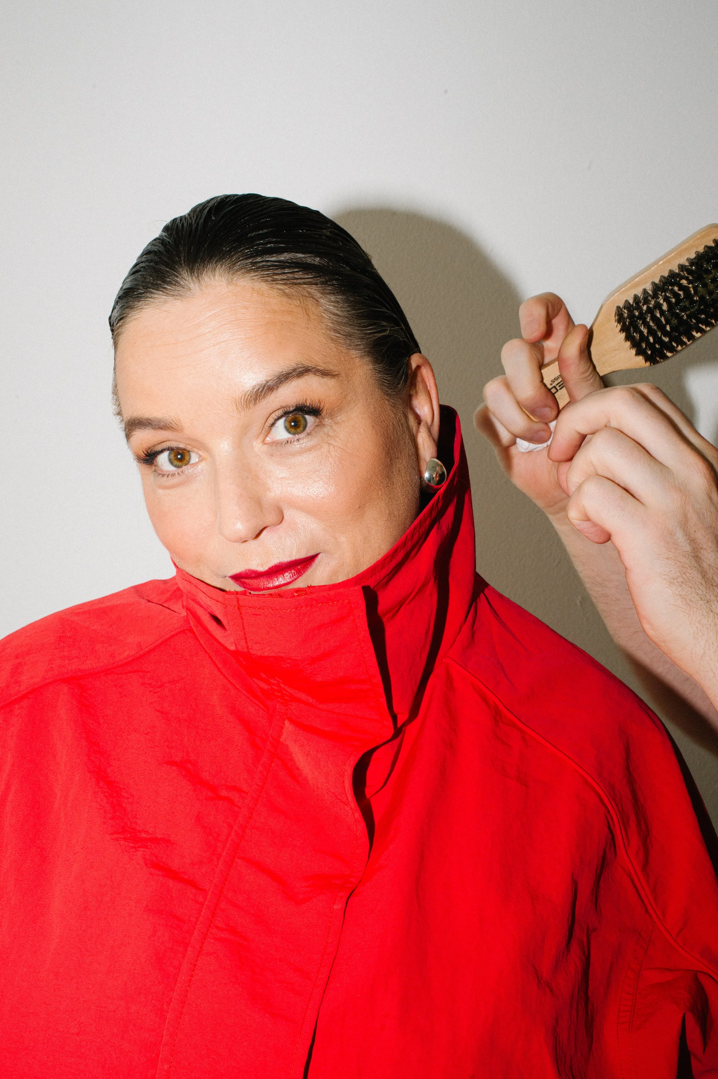 Woman with slicked-back dark hair, wearing a red jacket, stylist holding a hairbrush near her head, standing against a plain gray background.