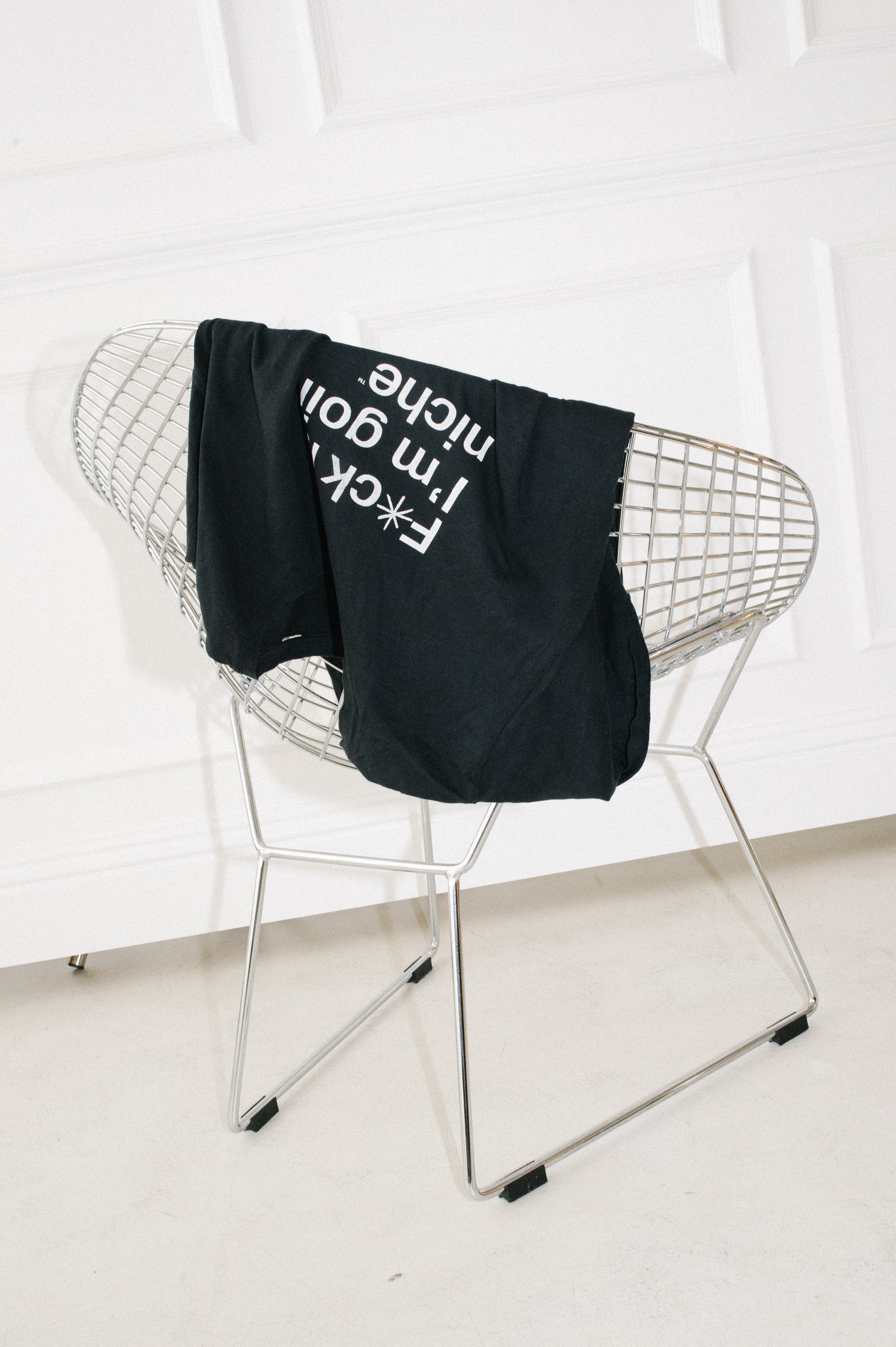 A metal wire chair with black iconic 'F*ck it I'm going niche'  tee draped over its backrest, placed against a white wall and white door.