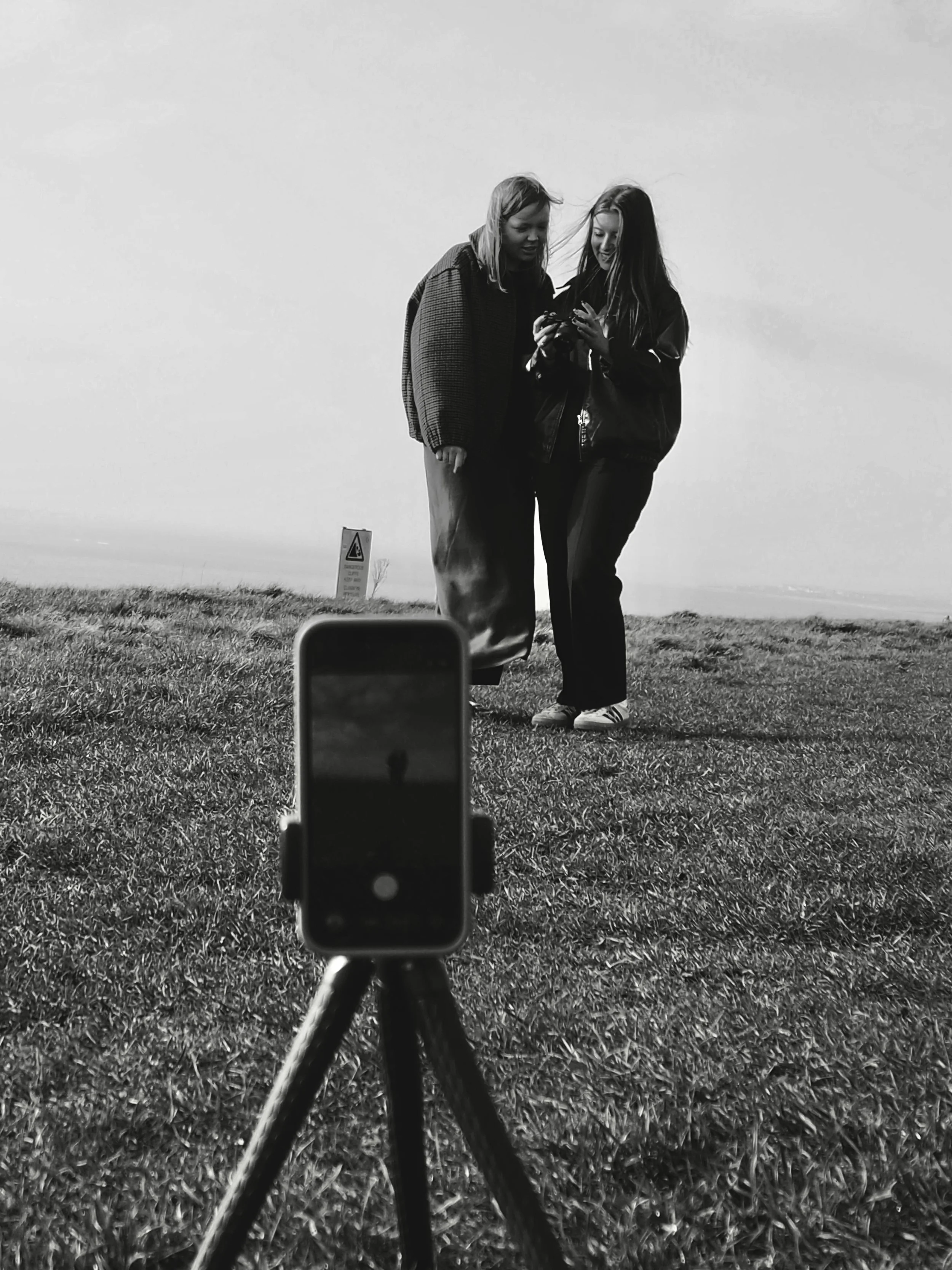 Two women are standing on a grassy field, looking at a smartphone together. A camera on a tripod in the foreground captures their photo. The scene is outdoors, with a sign visible in the background, and the photo is in black and white.