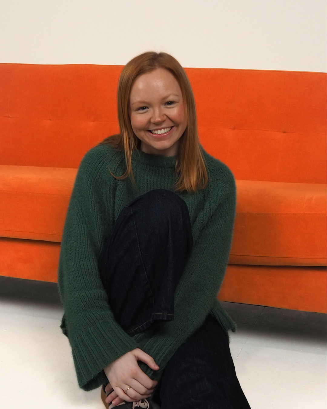 A young woman with red hair and a big smile, sitting on the floor in front of an orange couch, wearing a green sweater and black jeans.