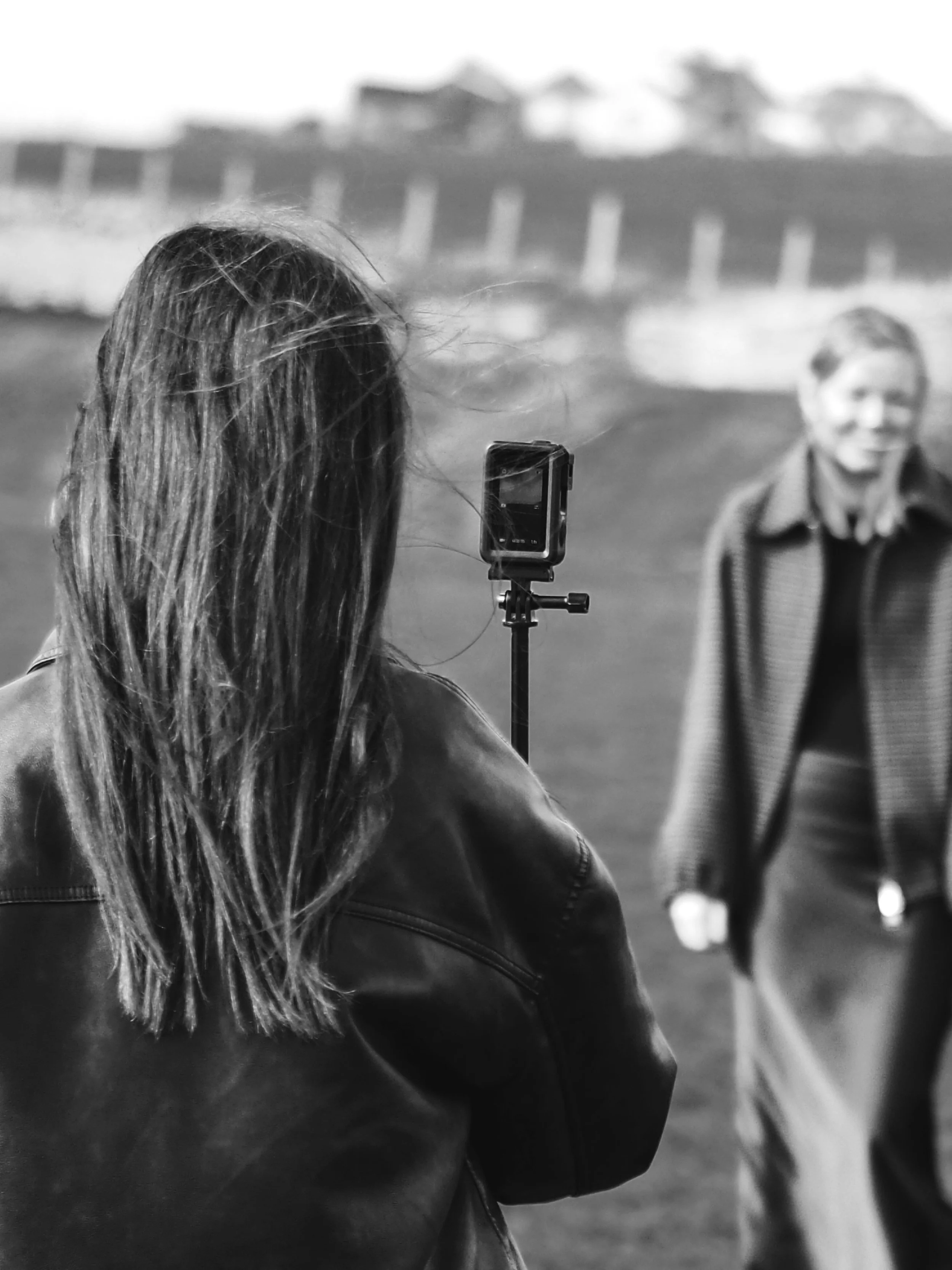 A woman with long hair wearing a leather jacket is taking a photo or recording a video of a smiling woman in a coat walking outdoors in a park or open space.