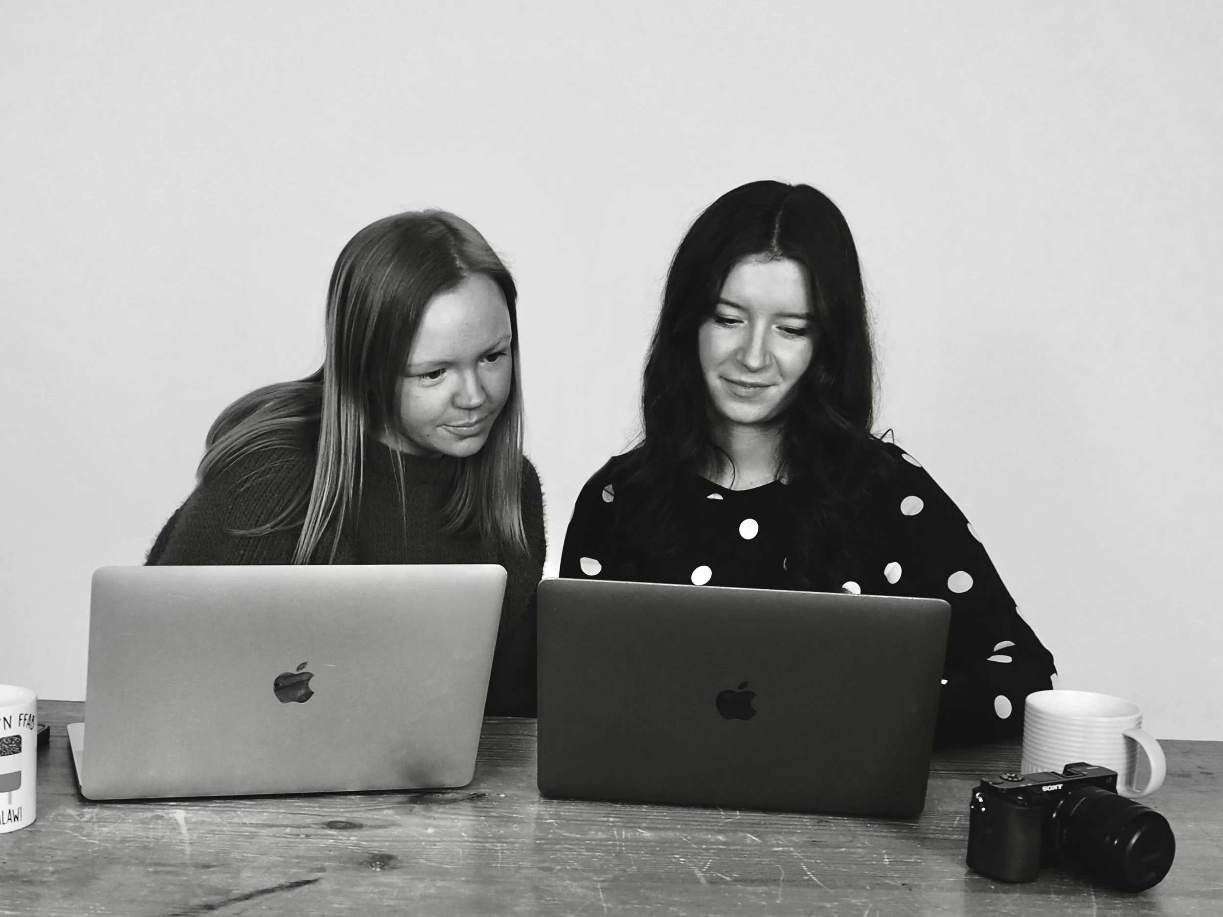 Two women sitting at a table with laptops, mugs, and a camera, looking at the screen together.