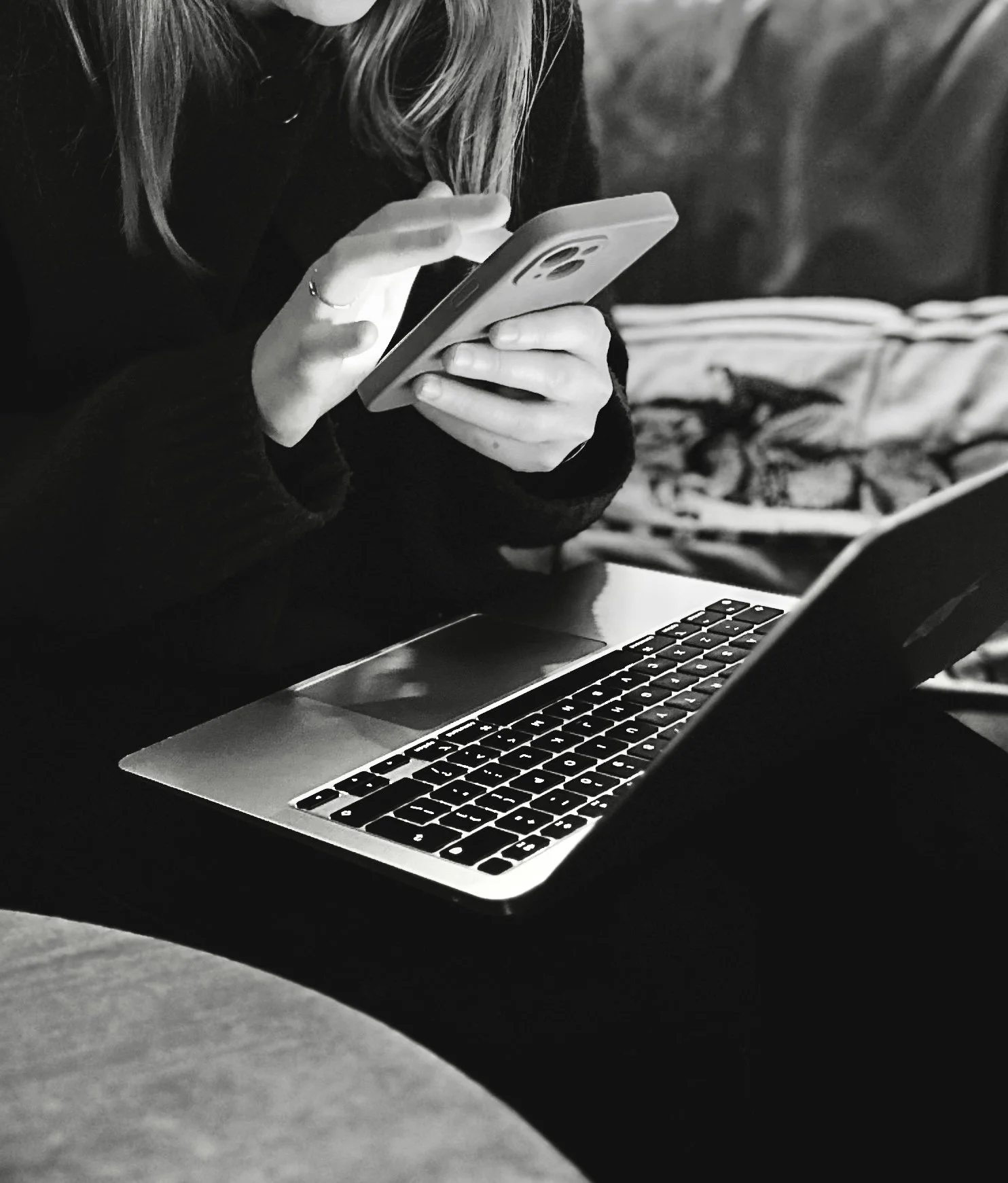 A person using a smartphone while sitting at a table with a laptop open in front. In the background, a dog is lying on a blanket.