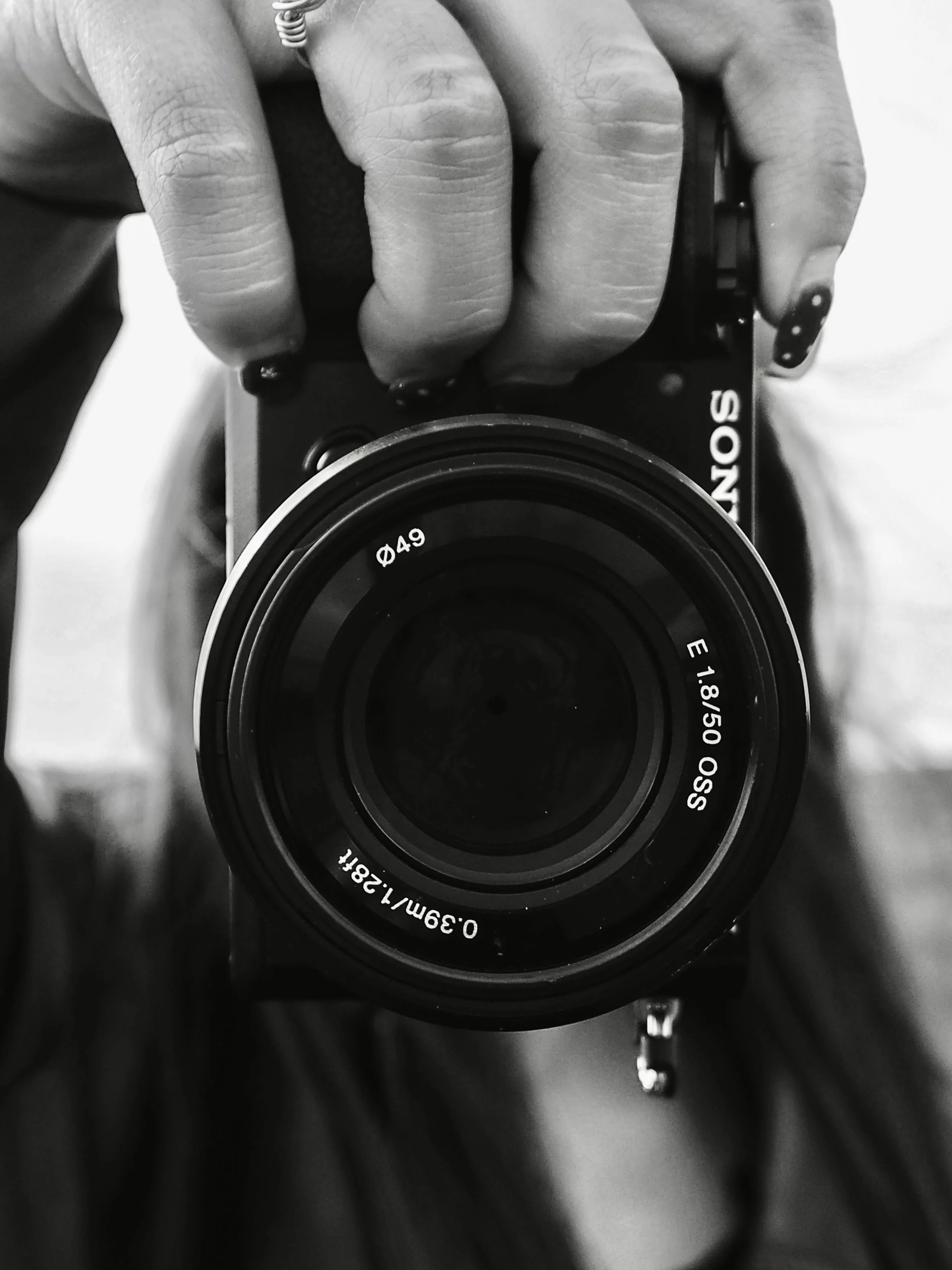 A person holding a Sony camera with an 18-50mm lens, visible fingers with painted nails, and a reflection of the camera lens in the photo.