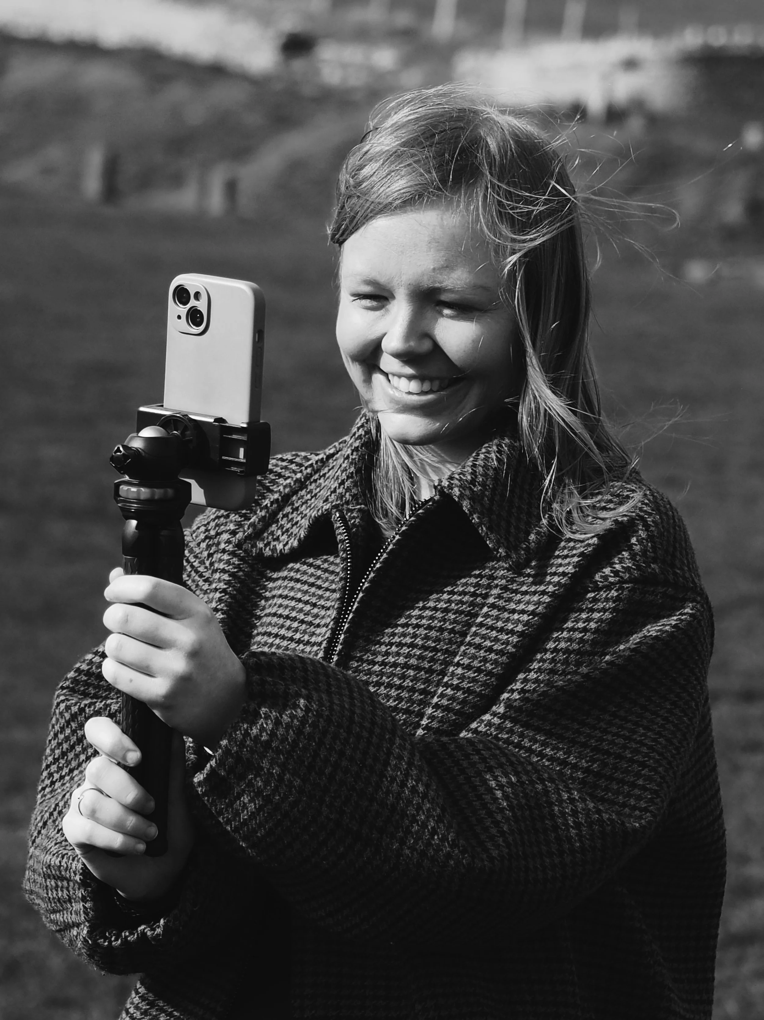 A young girl with a big smile takes a selfie outdoors with a smartphone mounted on a tripod.