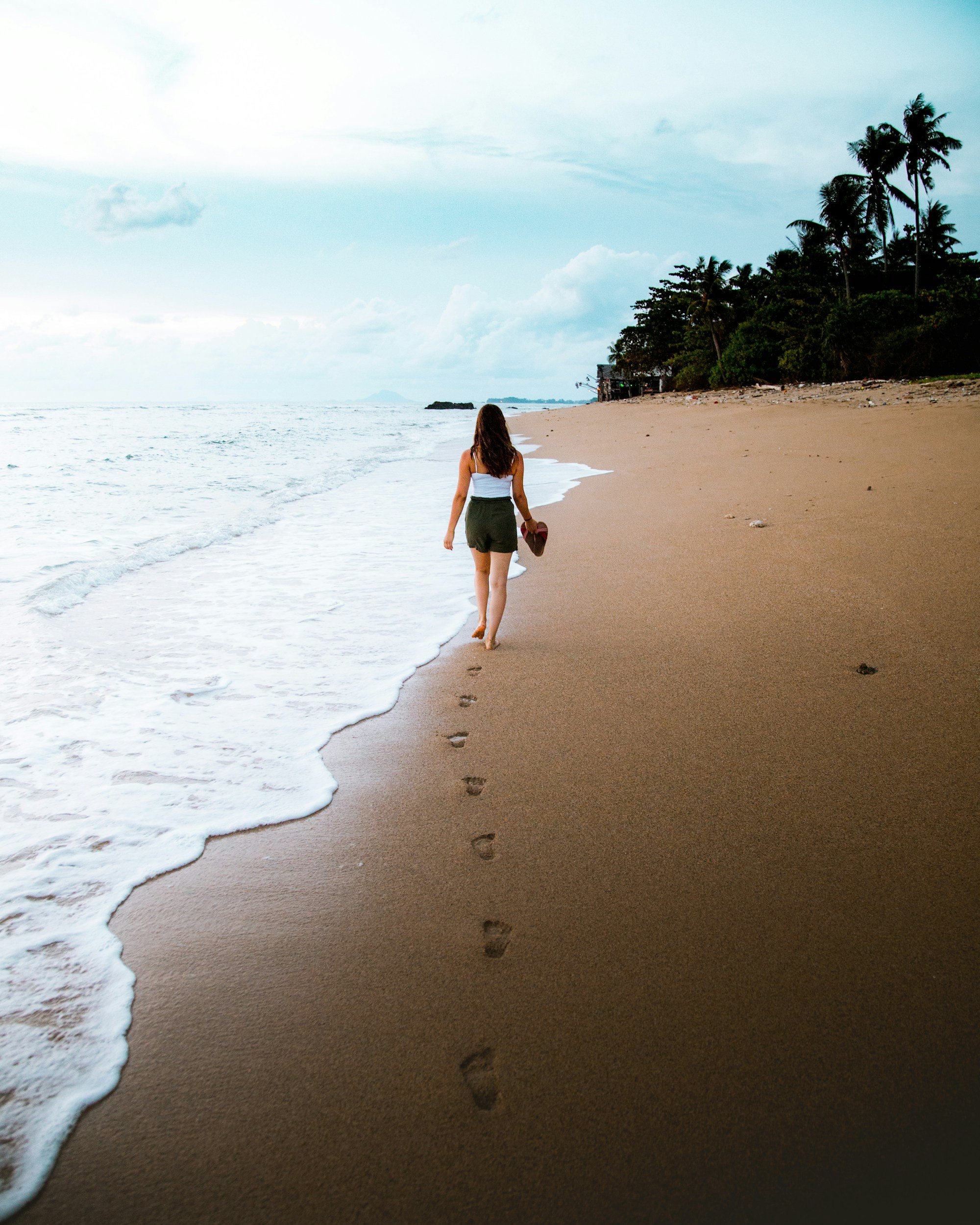 A woman walking on a sandy beach with footprints in the sand, leaving footprints behind, near the shoreline with gentle waves and cloudy sky.