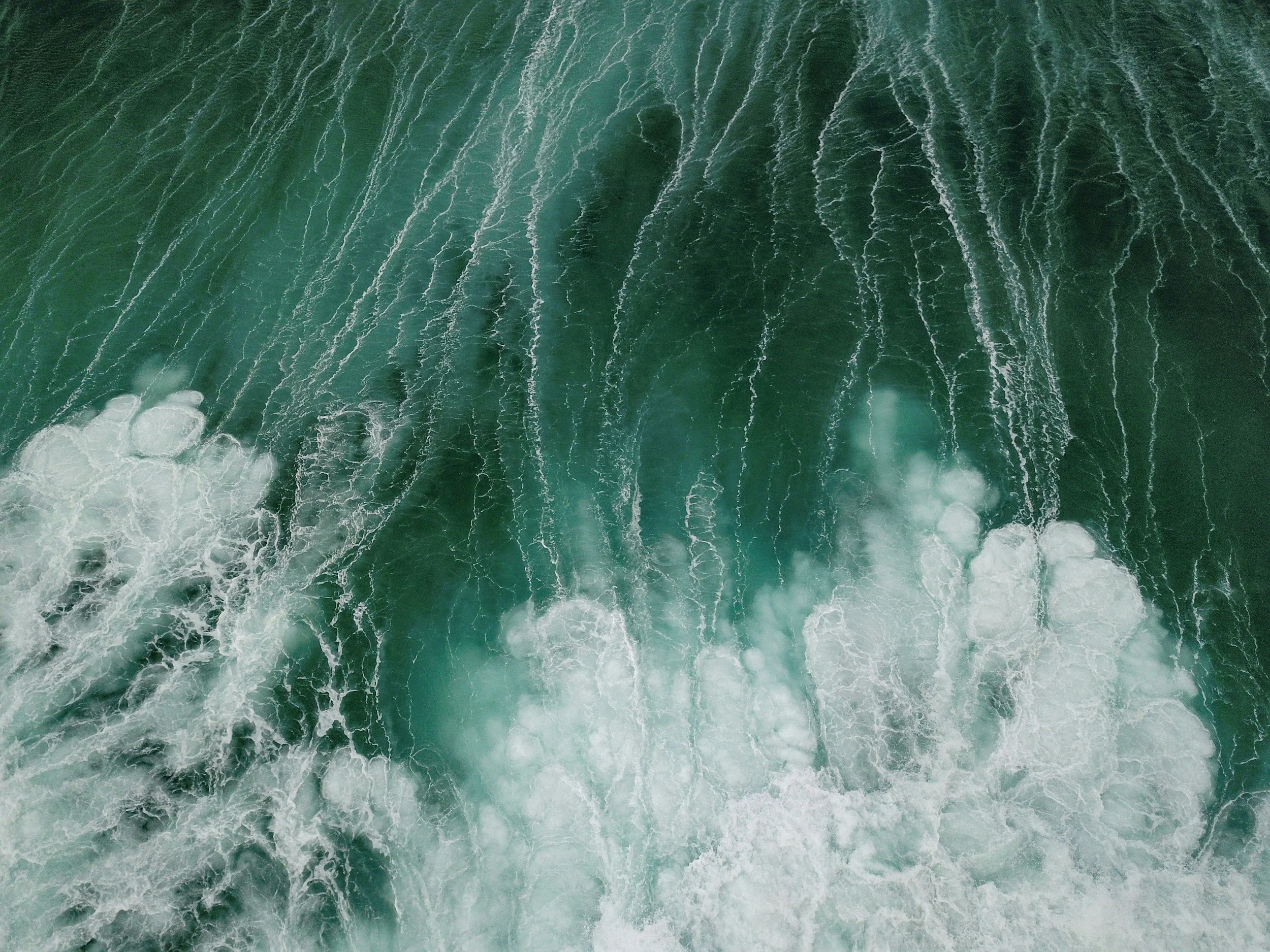 Aerial view of ocean waves breaking and creating white foam on teal water.