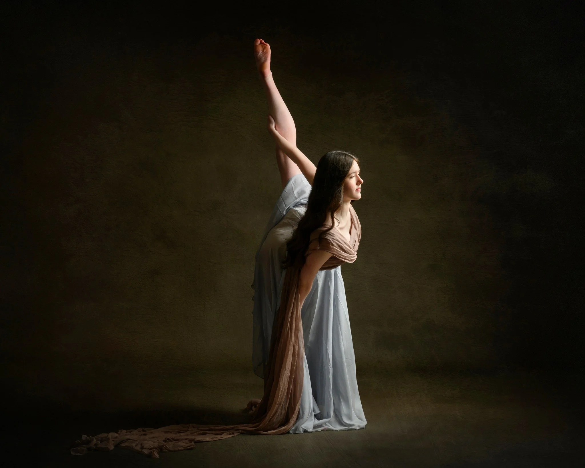 A woman in a white gown is doing a yoga pose with one arm raised and bent behind her back, with her eyes closed, in a dimly lit setting.