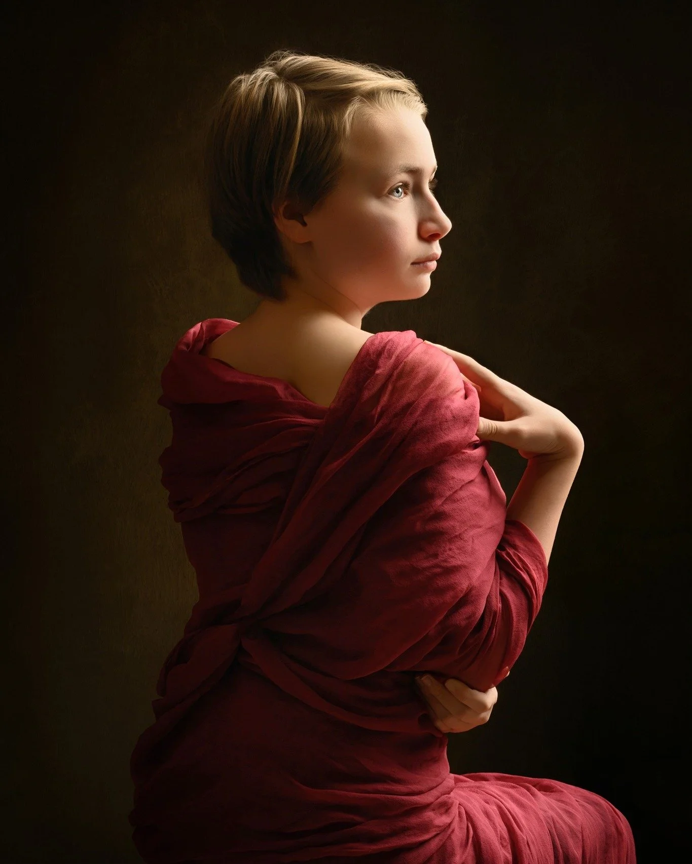 A woman with short, light brown hair, sitting in profile against a dark background, wearing a red shawl.
